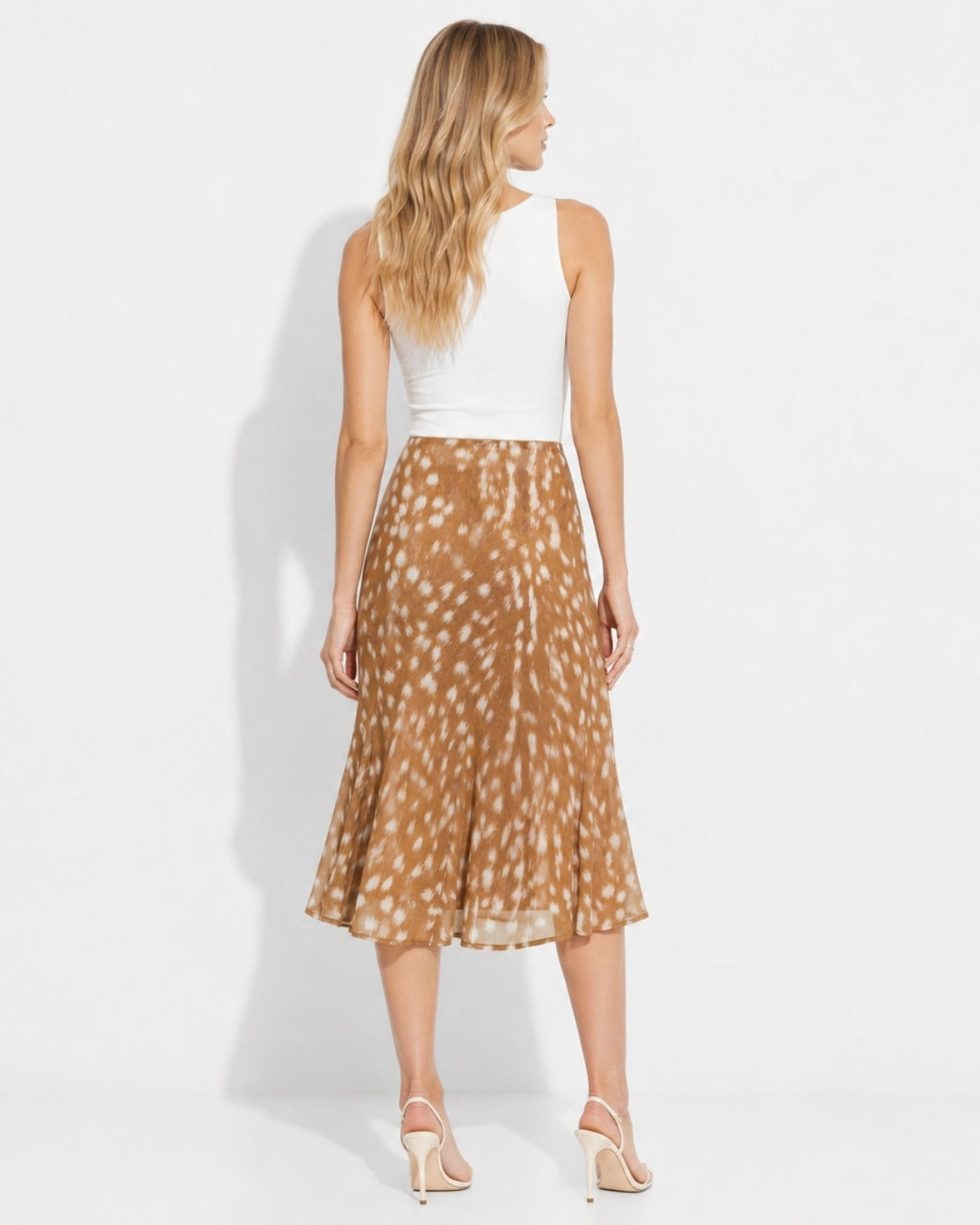 A woman shown from the back wearing a sleeveless white top and a brown midi skirt with a white spotted animal-like print, standing against a plain white studio background.