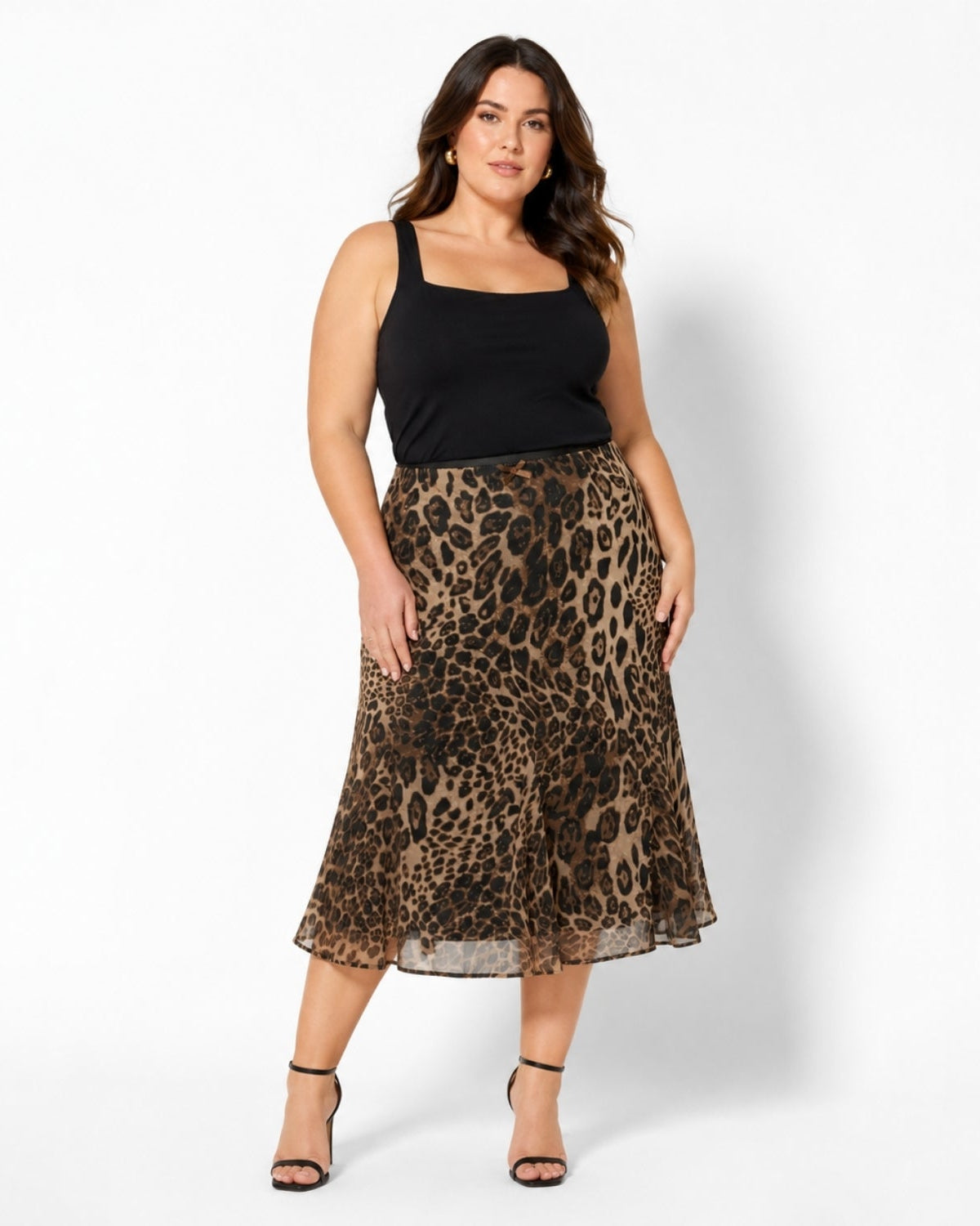 A woman modeling a leopard-print midi skirt with a black camisole top and black strappy heels, standing against a plain white studio background.