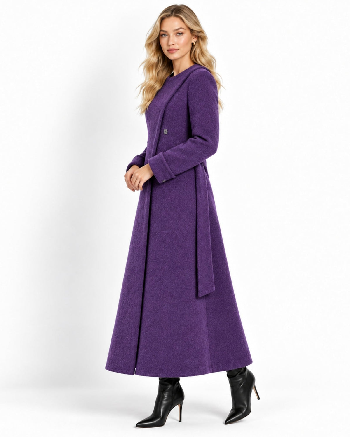 A woman modeling a long, double-breasted purple coat with a waist tie, paired with black pointed-toe ankle boots, against a plain white studio background.
