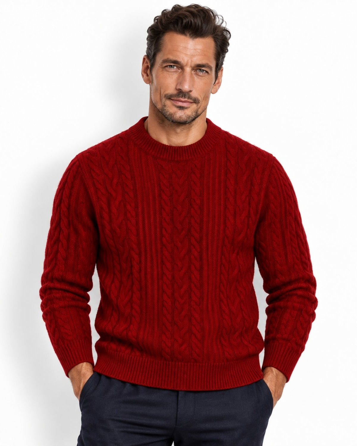 A man wearing a red cable-knit crewneck sweater and navy trousers, standing against a plain white studio background.