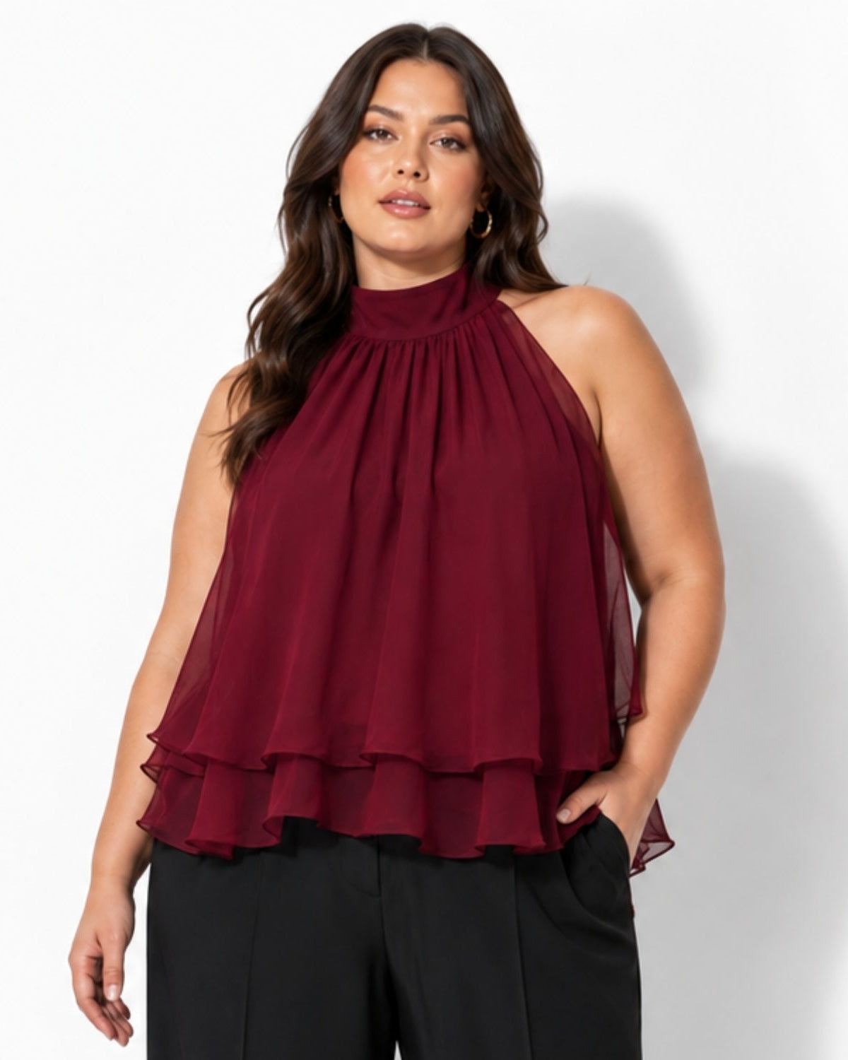 A woman modeling a sleeveless high-neck blouse in deep burgundy with a gathered, pleated ruffled hem, paired with black pants, against a plain white studio background.