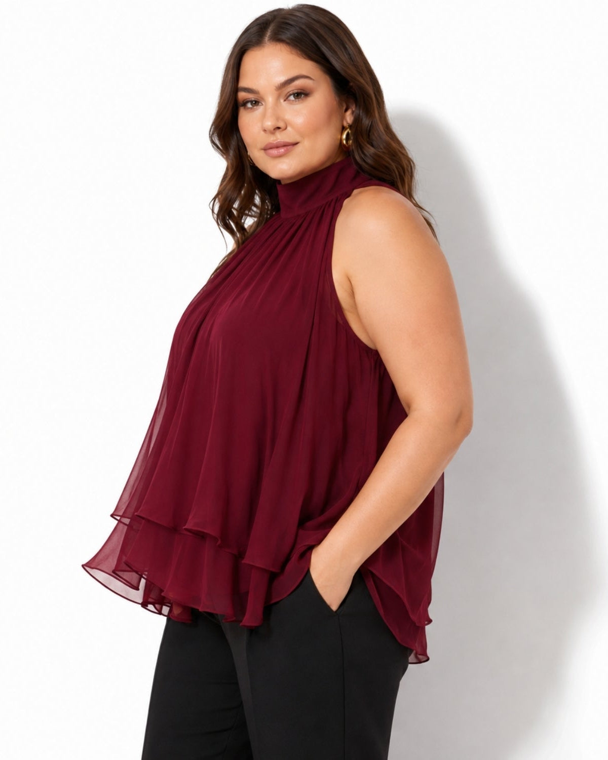 A woman wearing a sleeveless high-neck pleated blouse in deep burgundy, paired with black pants, standing against a white studio background.
