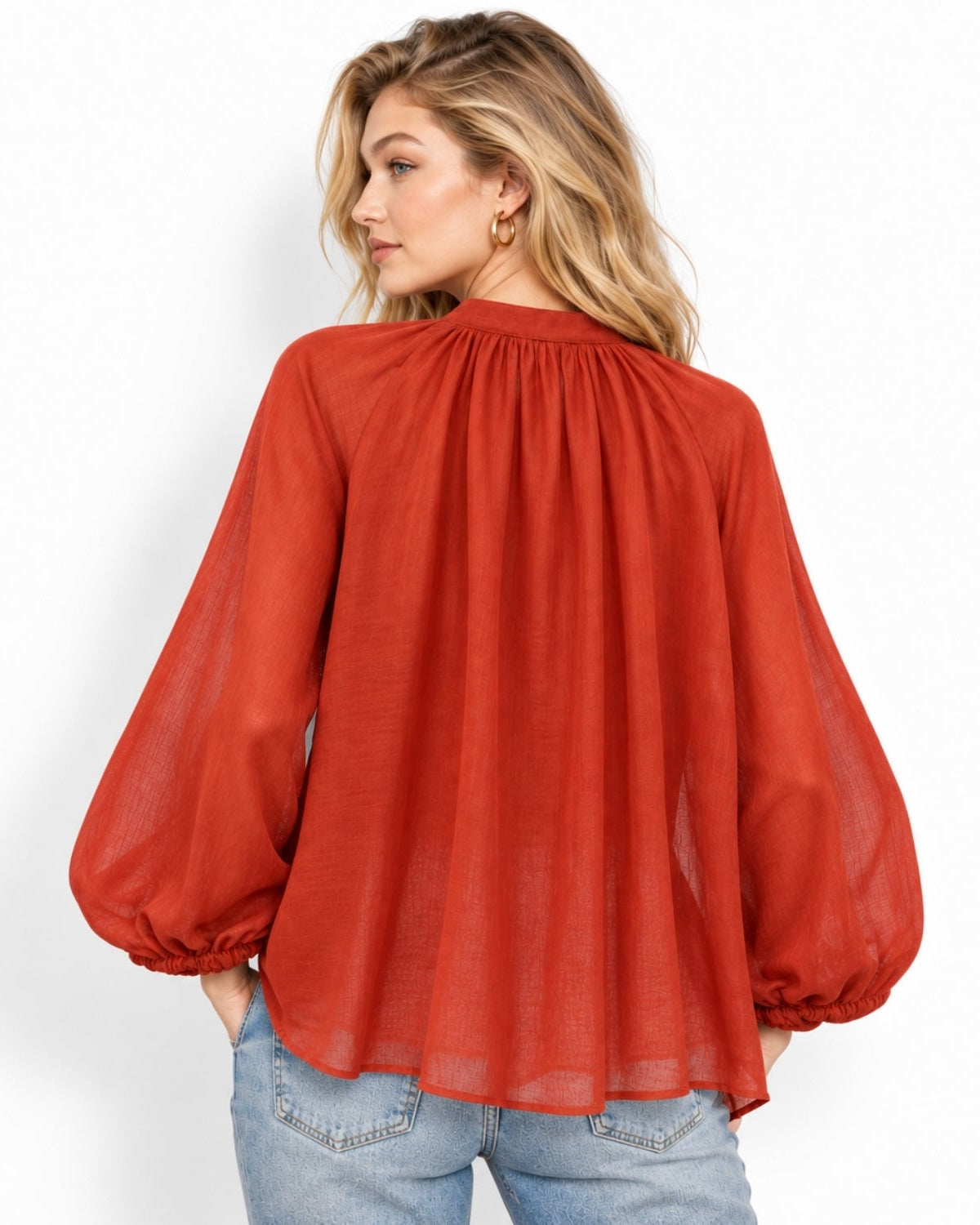 A woman wearing a rust-orange long-sleeve blouse with a gathered, balloon-style back, paired with light-wash blue jeans, shown from a three-quarters back view against a white studio background.