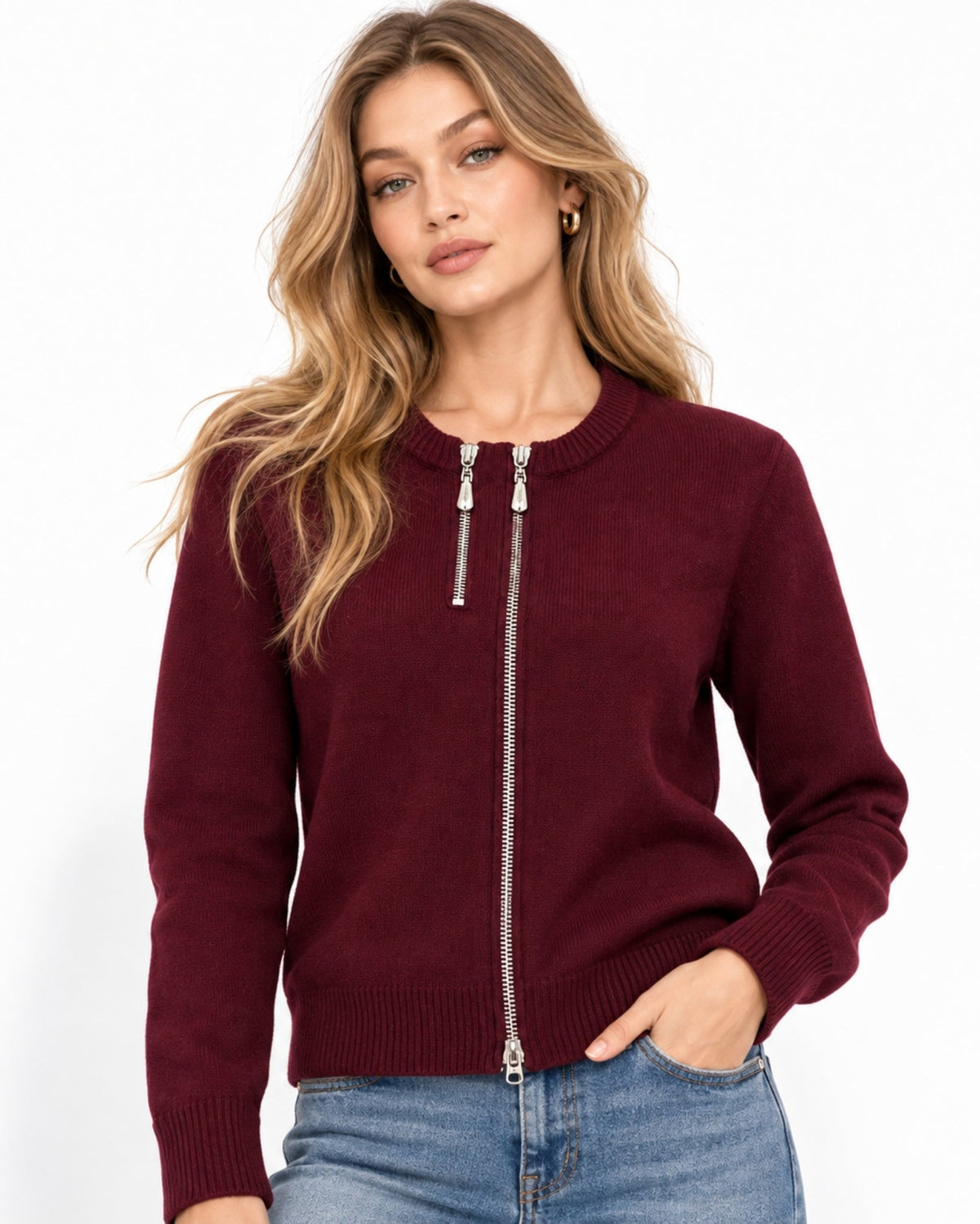 A woman wearing a burgundy knit zip-up sweater with a silver zipper and blue jeans, photographed against a clean white studio background.