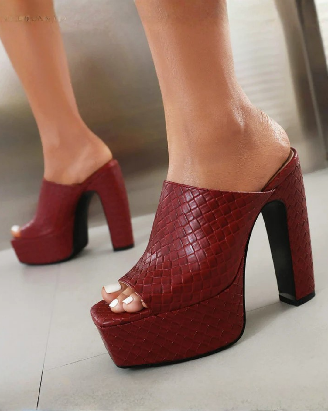 A pair of red textured platform mule heels with high block heels, shown on a model's feet against a neutral indoor background.