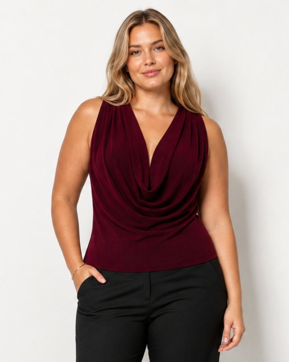 A woman modeling a sleeveless draped cowl-neck top in deep burgundy, paired with black pants, against a plain light gray studio background.