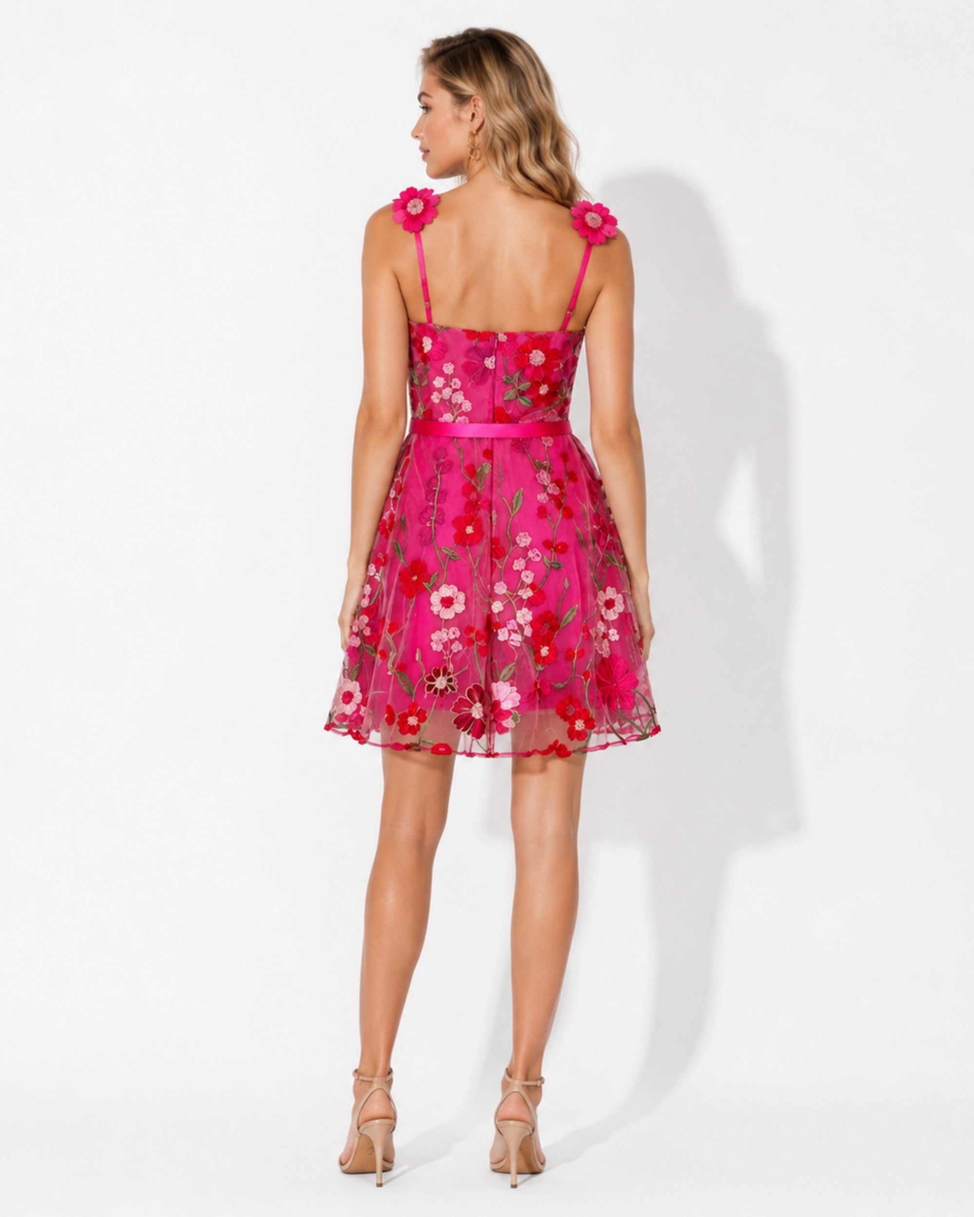 A woman modeling a short pink floral dress from the back, featuring floral embroidery, thin straps, and flower embellishments on the shoulders, set against a plain white studio background.