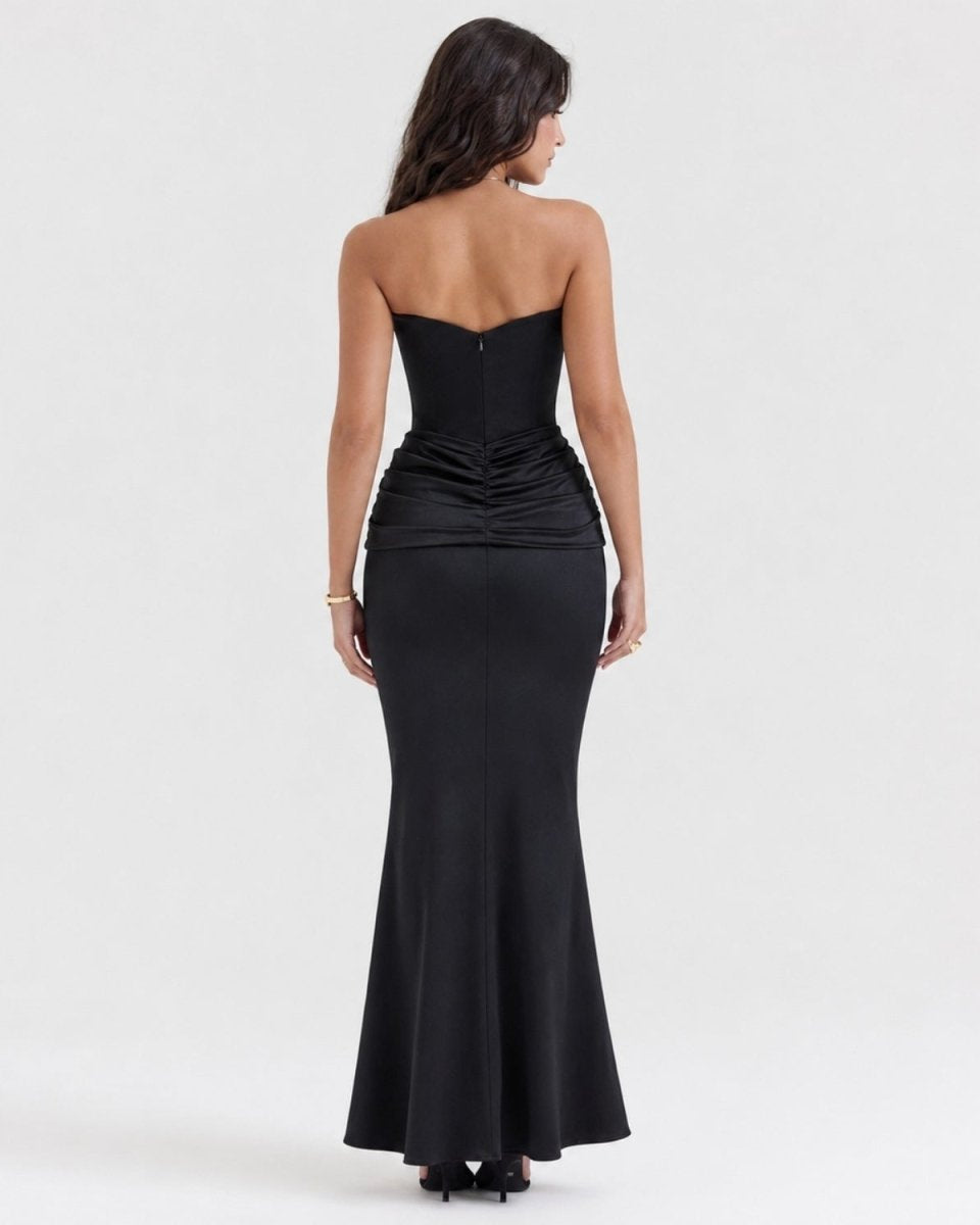 A woman viewed from the back wearing a strapless black formal gown with a structured corset-style bodice, gathered waist, and fitted mermaid-style skirt on a light gray studio background.