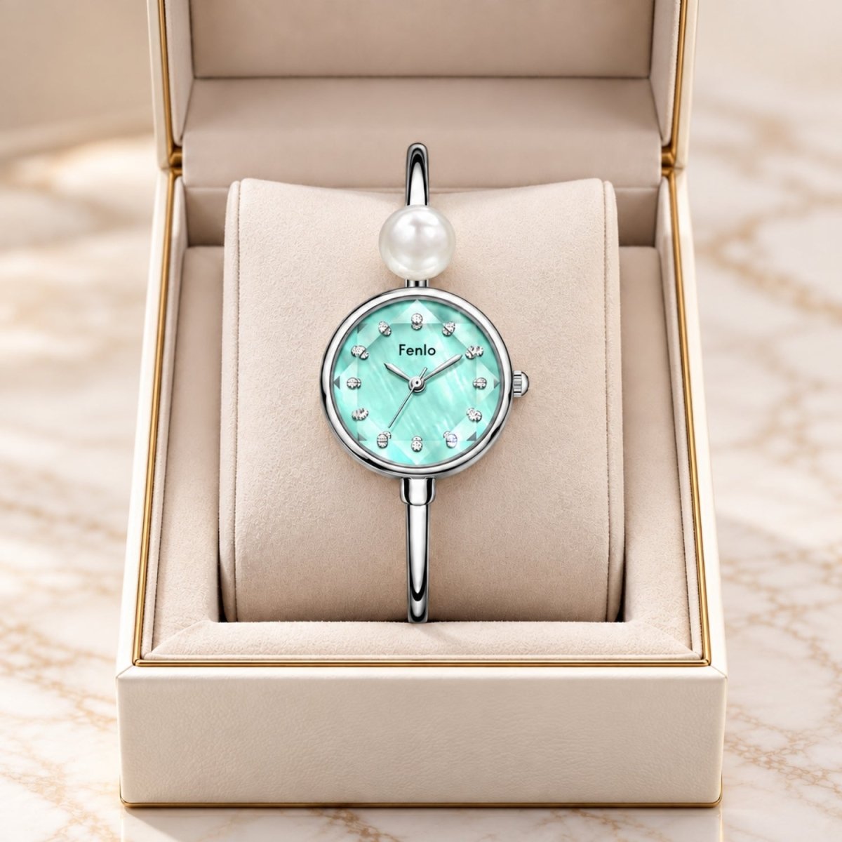 A silver-tone bangle watch with a round mint green dial, crystal hour markers, and a single white pearl at the top, displayed inside an open beige jewelry box on a marble surface.