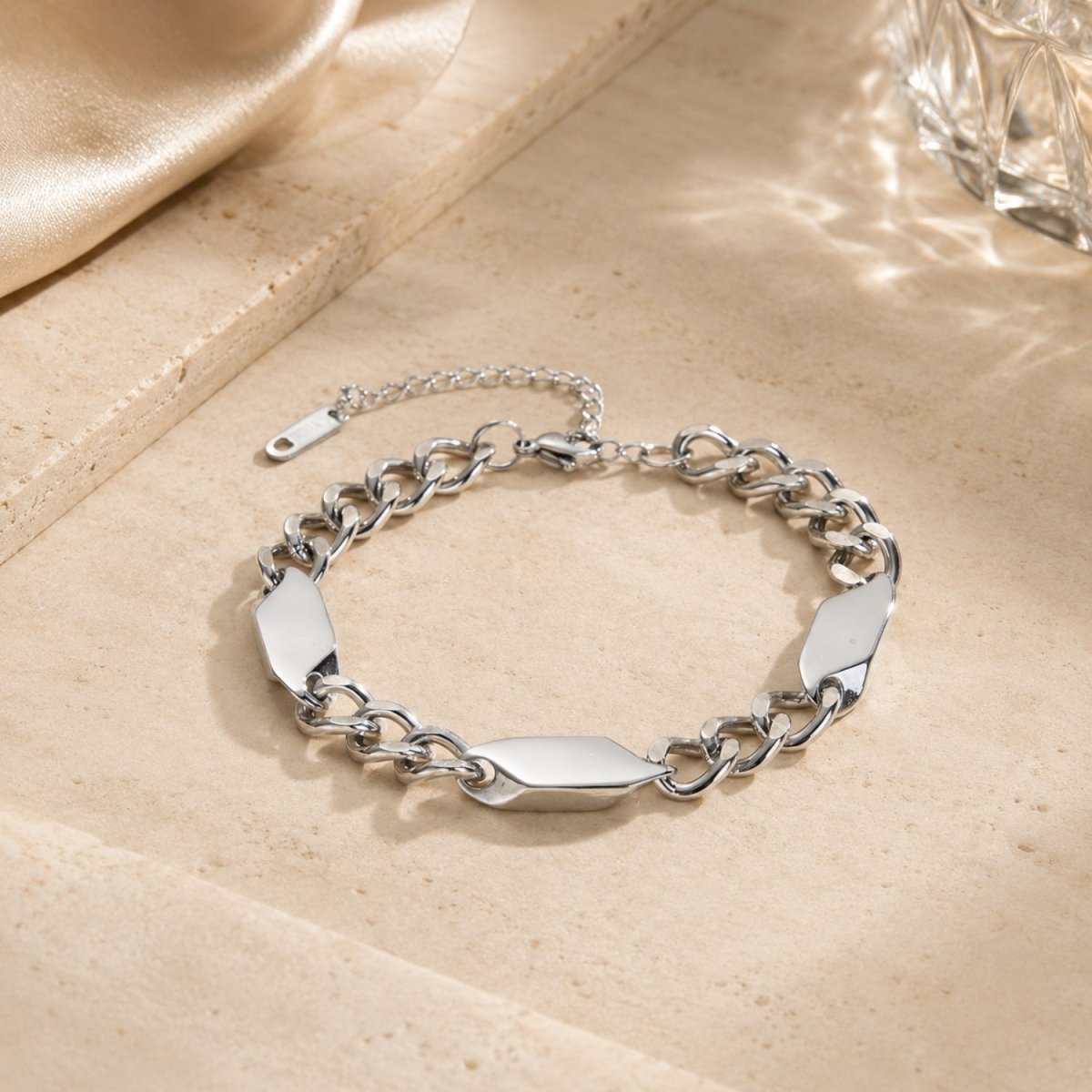 Silver stainless steel geometric link bracelet laid flat on a textured beige stone surface in warm sunlight