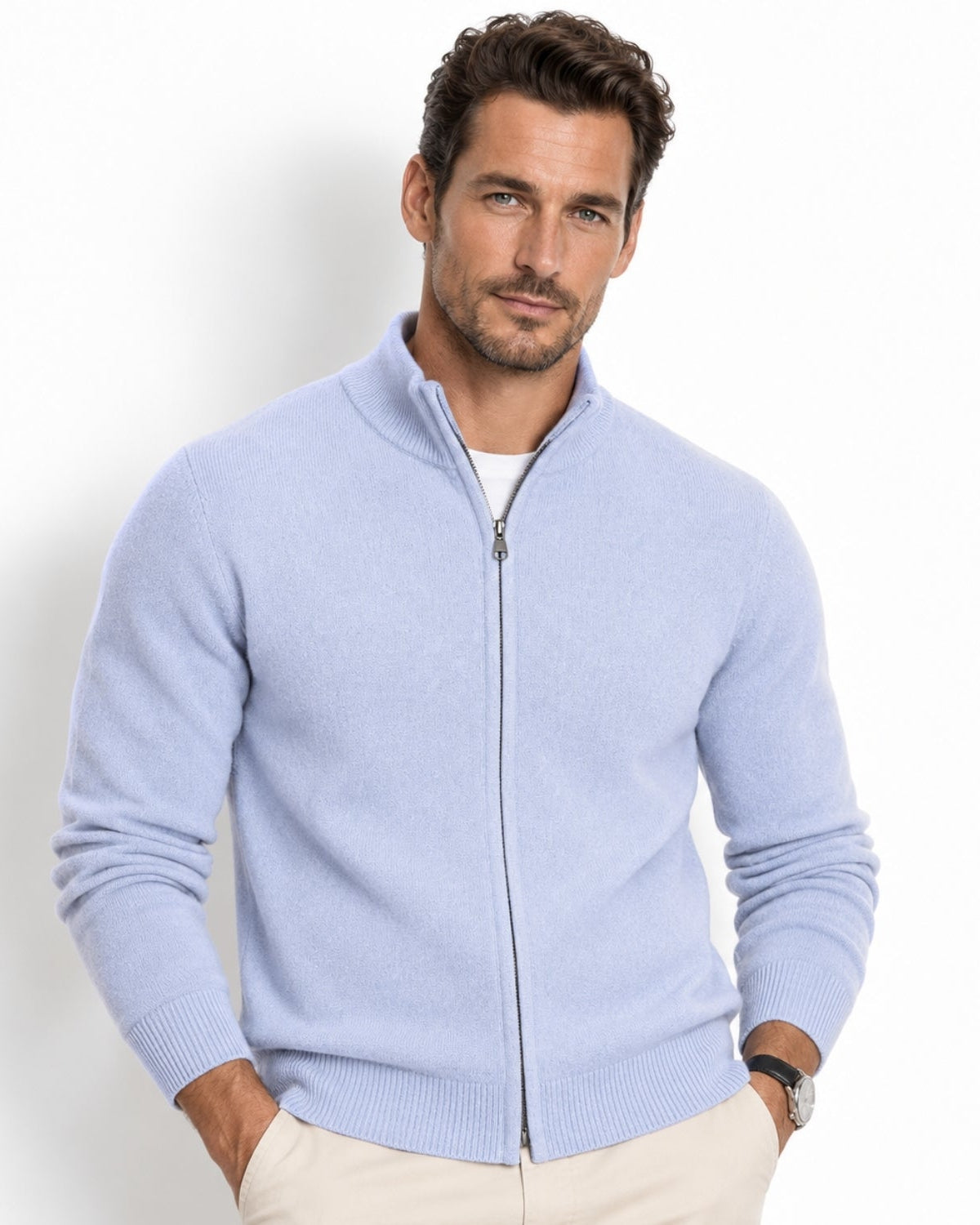 A man wearing a sky blue zip-up knit sweater over a white undershirt and beige pants, standing against a plain white studio background.