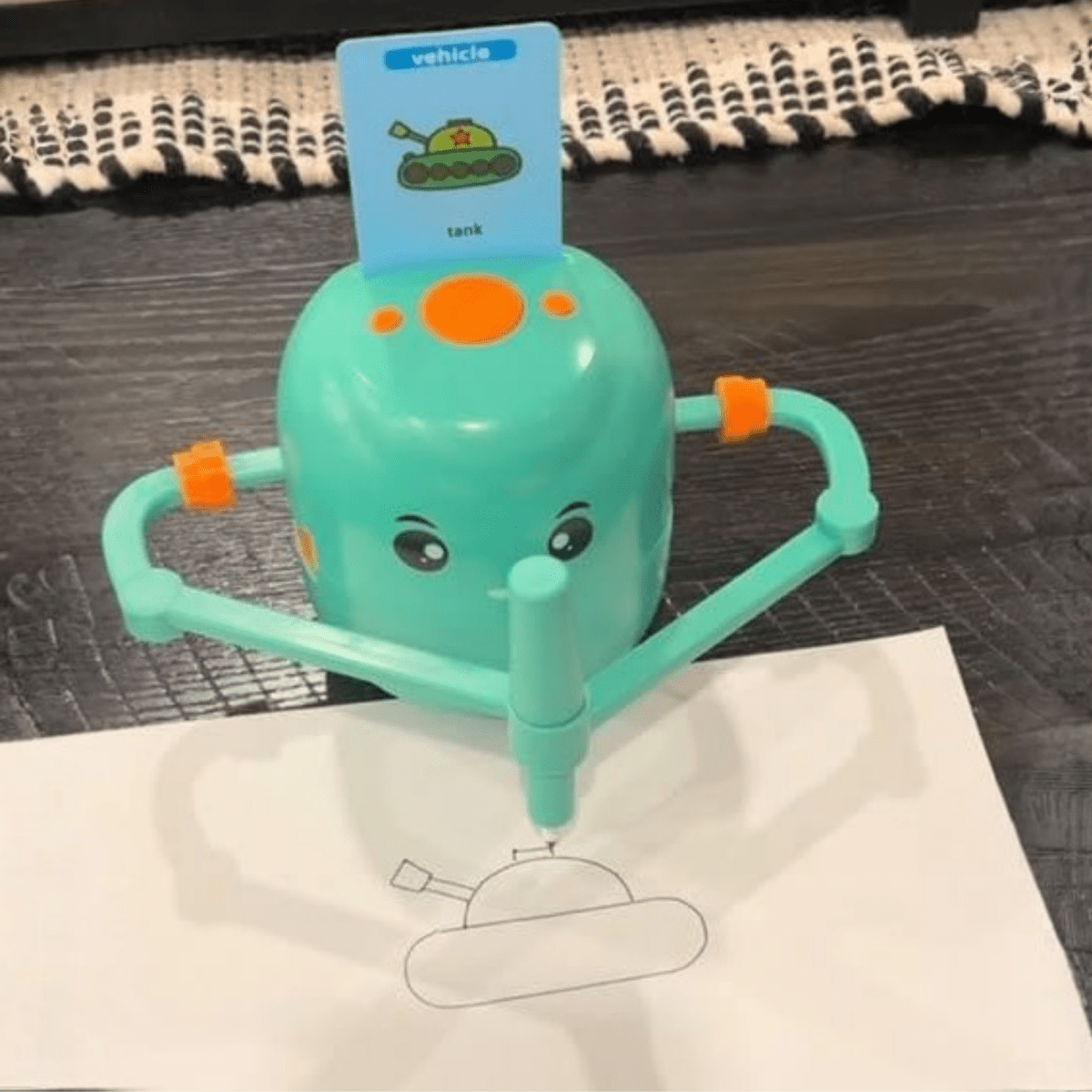 A teal drawing toy for kids with a cartoon face, featuring arm-like supports and a card labeled 'vehicle' and 'tank' propped behind it, drawing on a white sheet of paper over a dark wood surface.