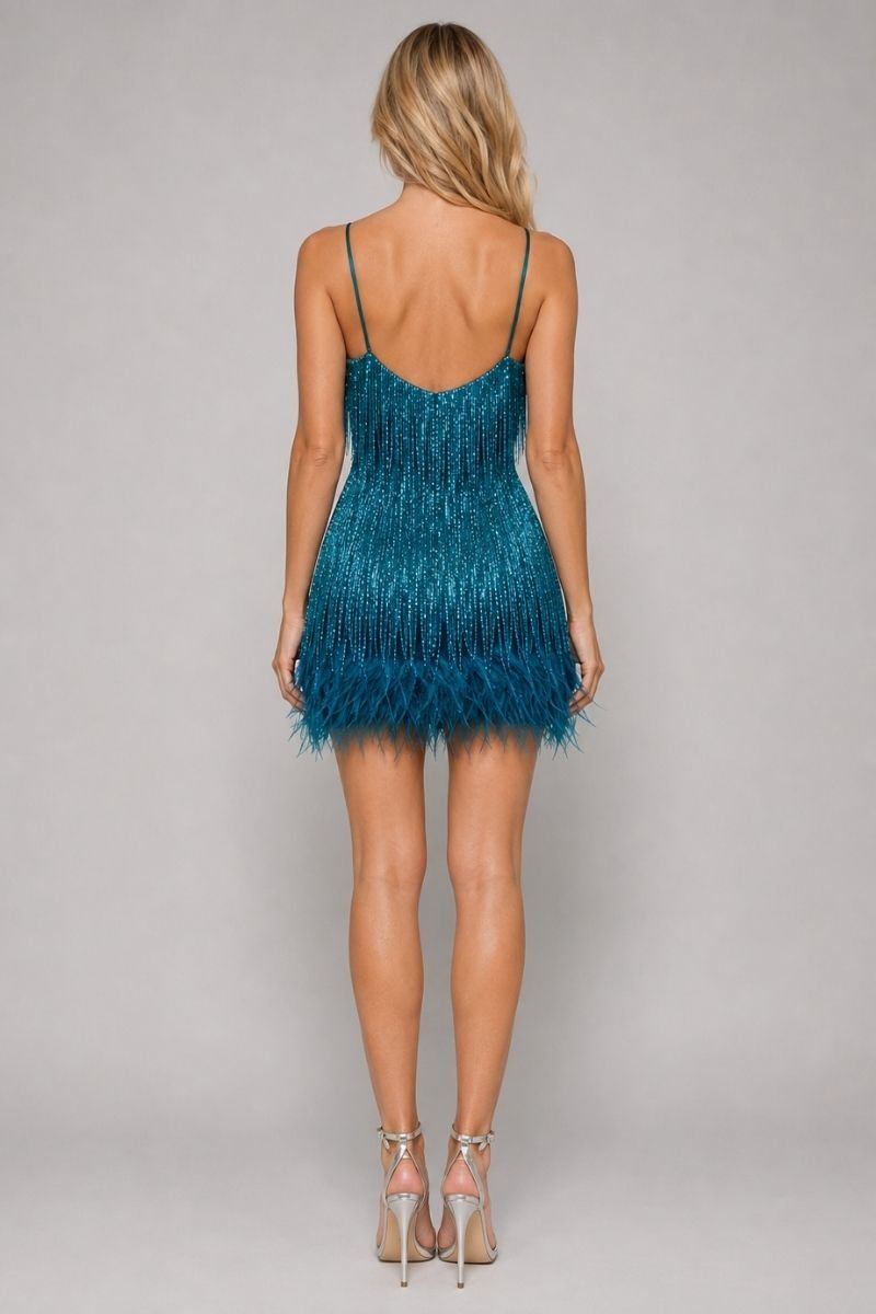A woman viewed from the back wearing a short teal fringe dress with thin straps and a low open back, paired with clear high-heeled sandals, against a light gray studio background.