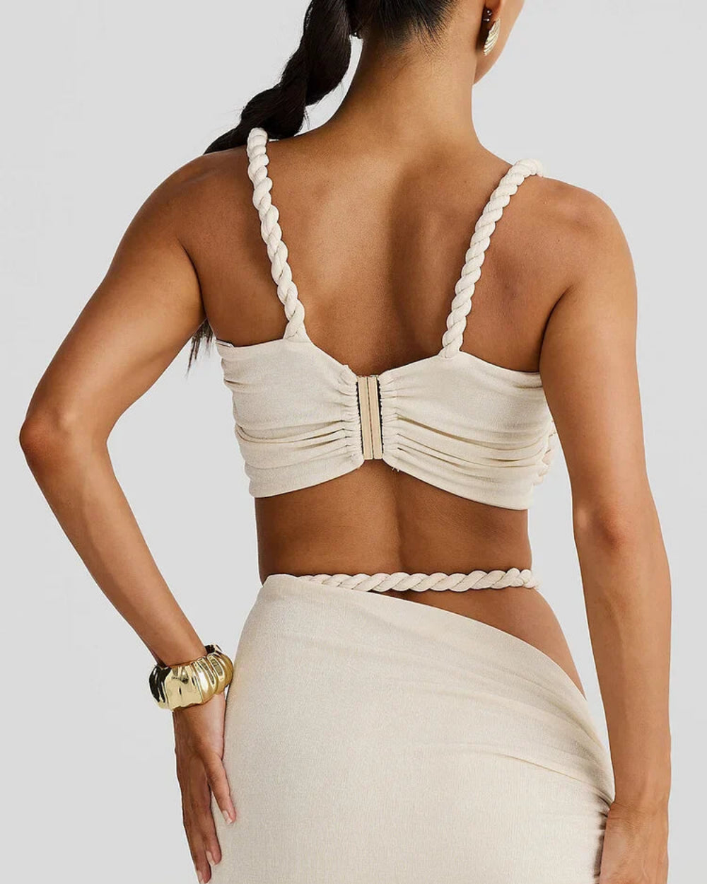 Model wearing The Thelma Set, a beige two-piece Grecian outfit featuring a rope detail bralette top and a ruched asymmetrical maxi skirt