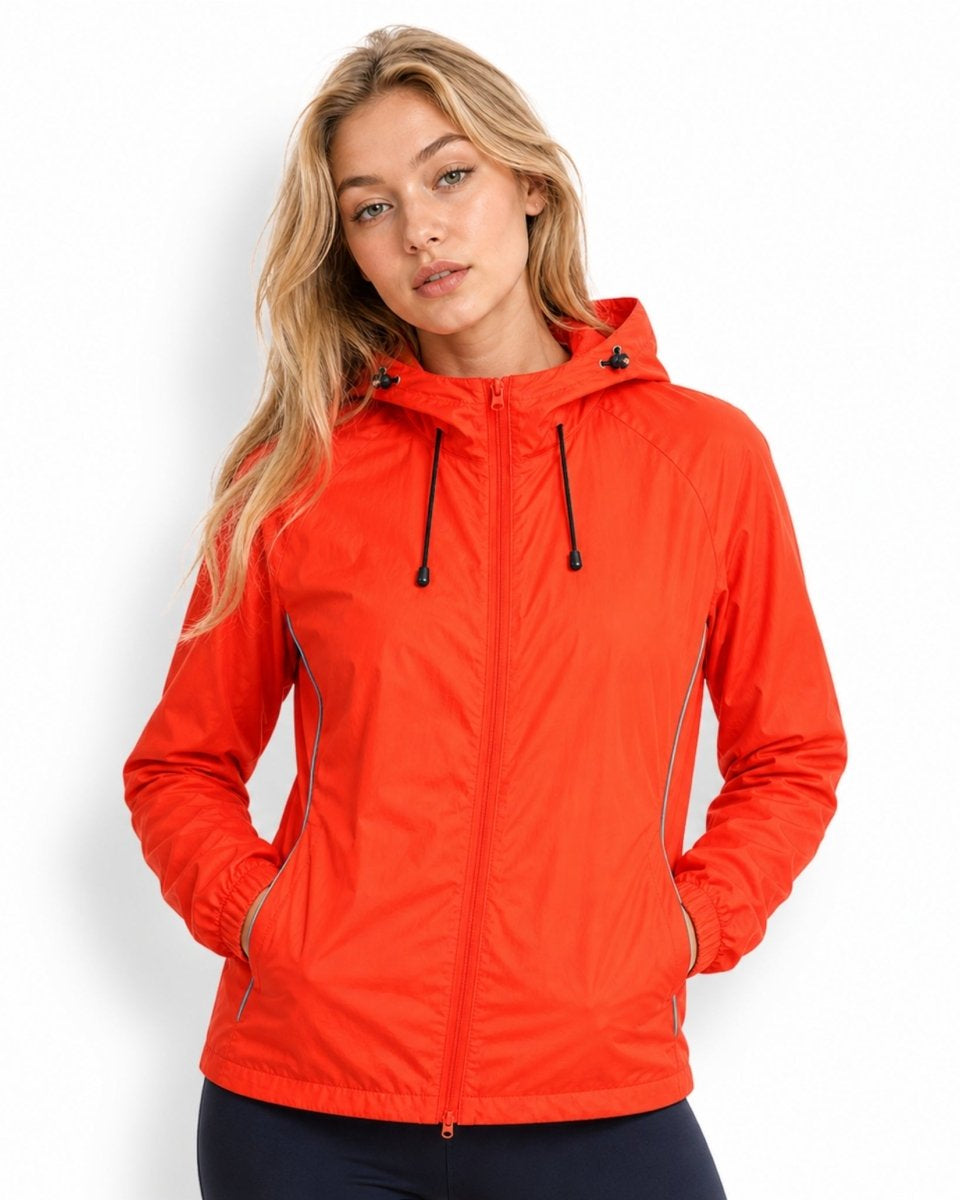 Blonde female model wearing bright orange lightweight hooded windbreaker jacket with hands casually resting in front pockets.