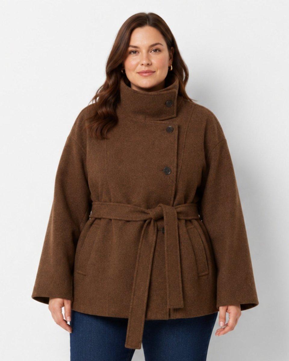 A woman modeling a brown wool-blend coat with a high mock neck, dark buttons, and a matching waist tie, paired with blue jeans against a light gray studio background.
