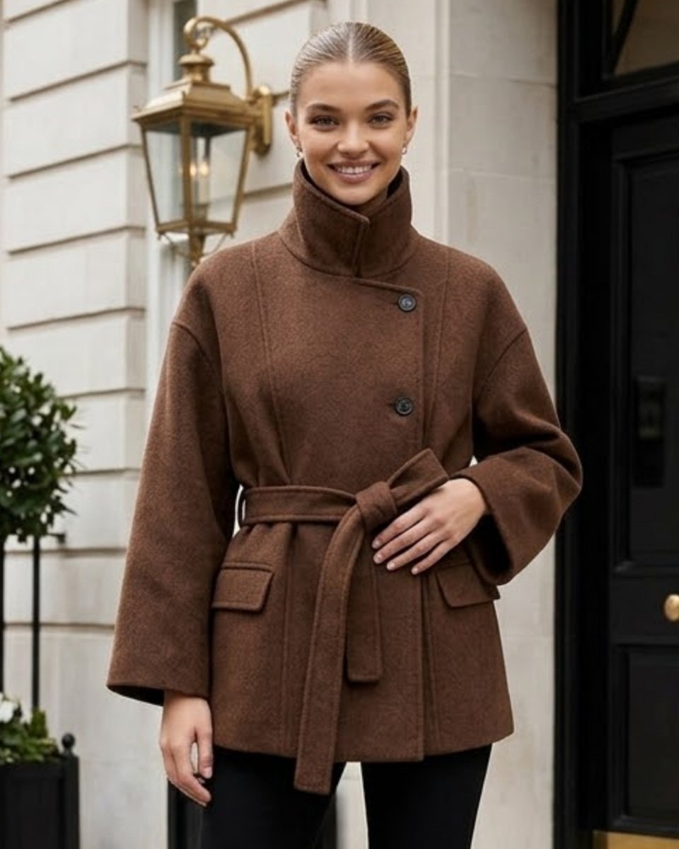 A woman wearing a brown belted coat with a high mock neck, standing in front of a white stone building exterior.