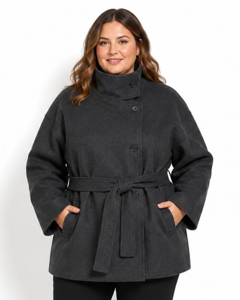 A woman modeling a dark charcoal gray belted coat with a high mock neck and front buttons, standing against a plain white background.