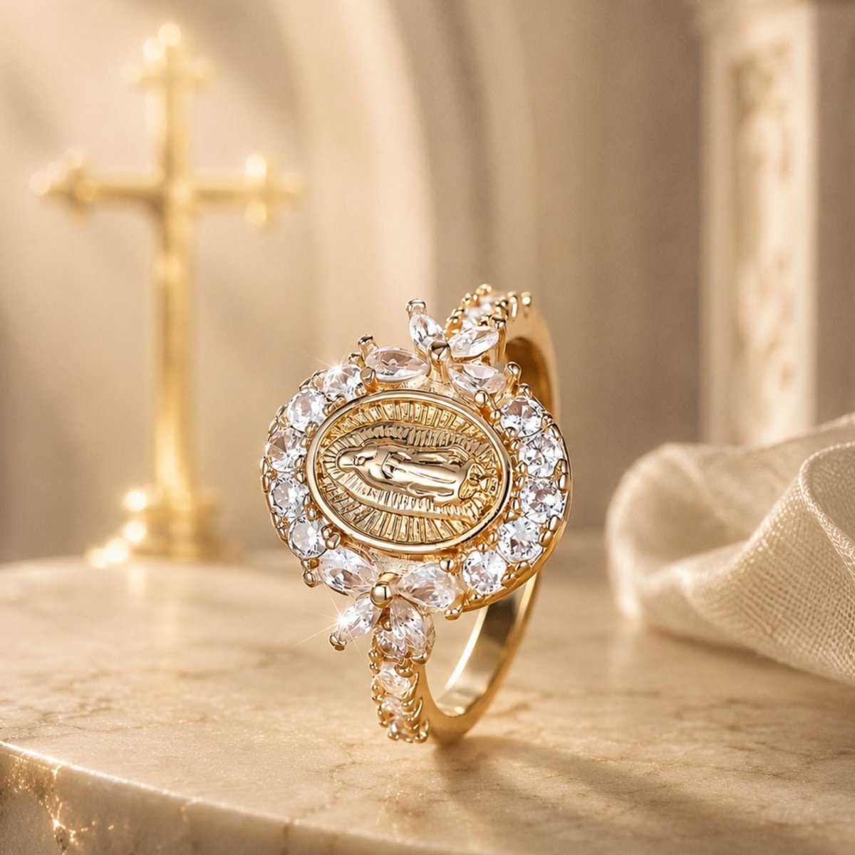A gold-toned religious ring featuring a Miraculous Medal design surrounded by clear crystals, displayed on a marble surface with a softly blurred gold cross in the background.