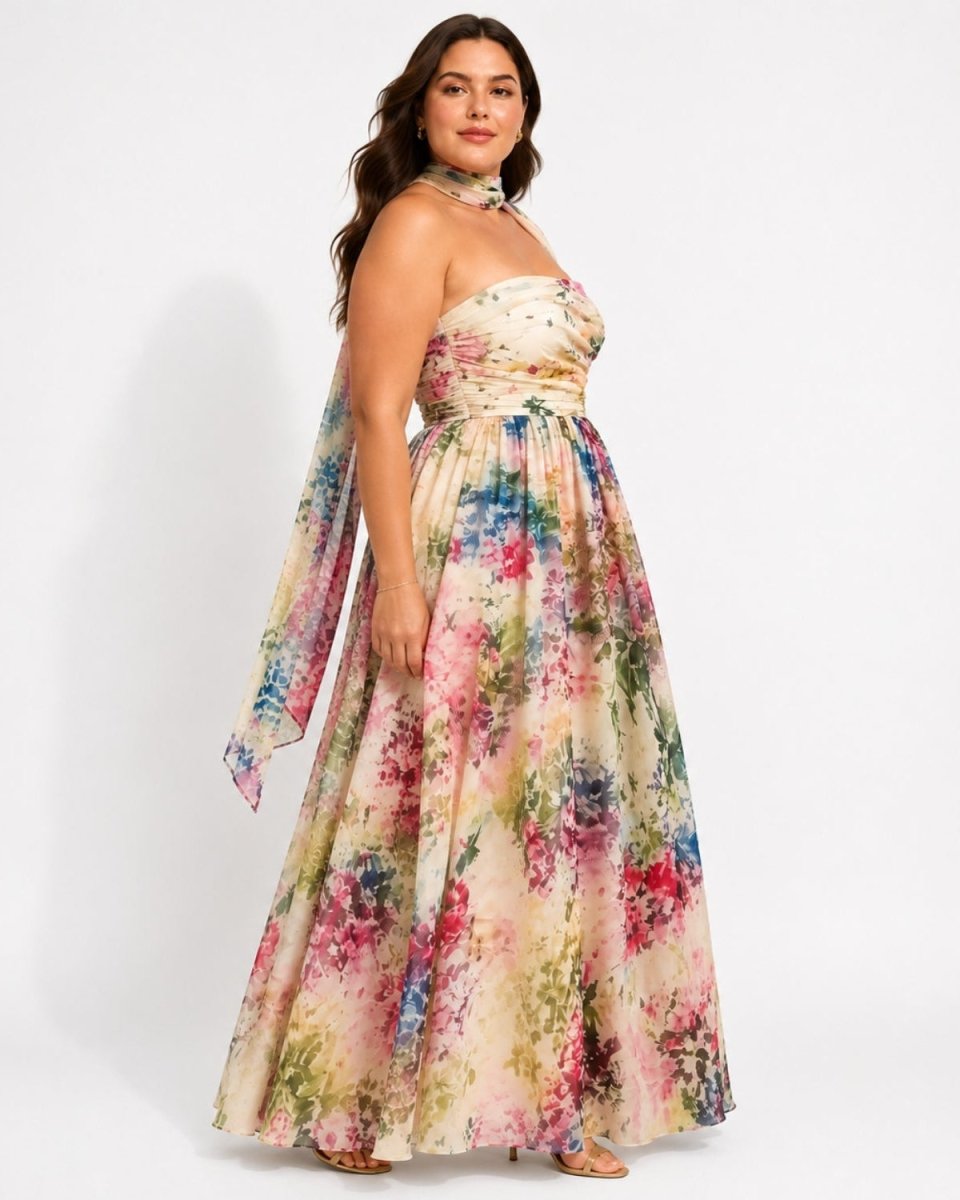 A woman models a strapless maxi dress with a floral watercolor print in pink, blue, green, and beige tones, featuring a high neck and a flowing skirt, set against a plain white studio background.