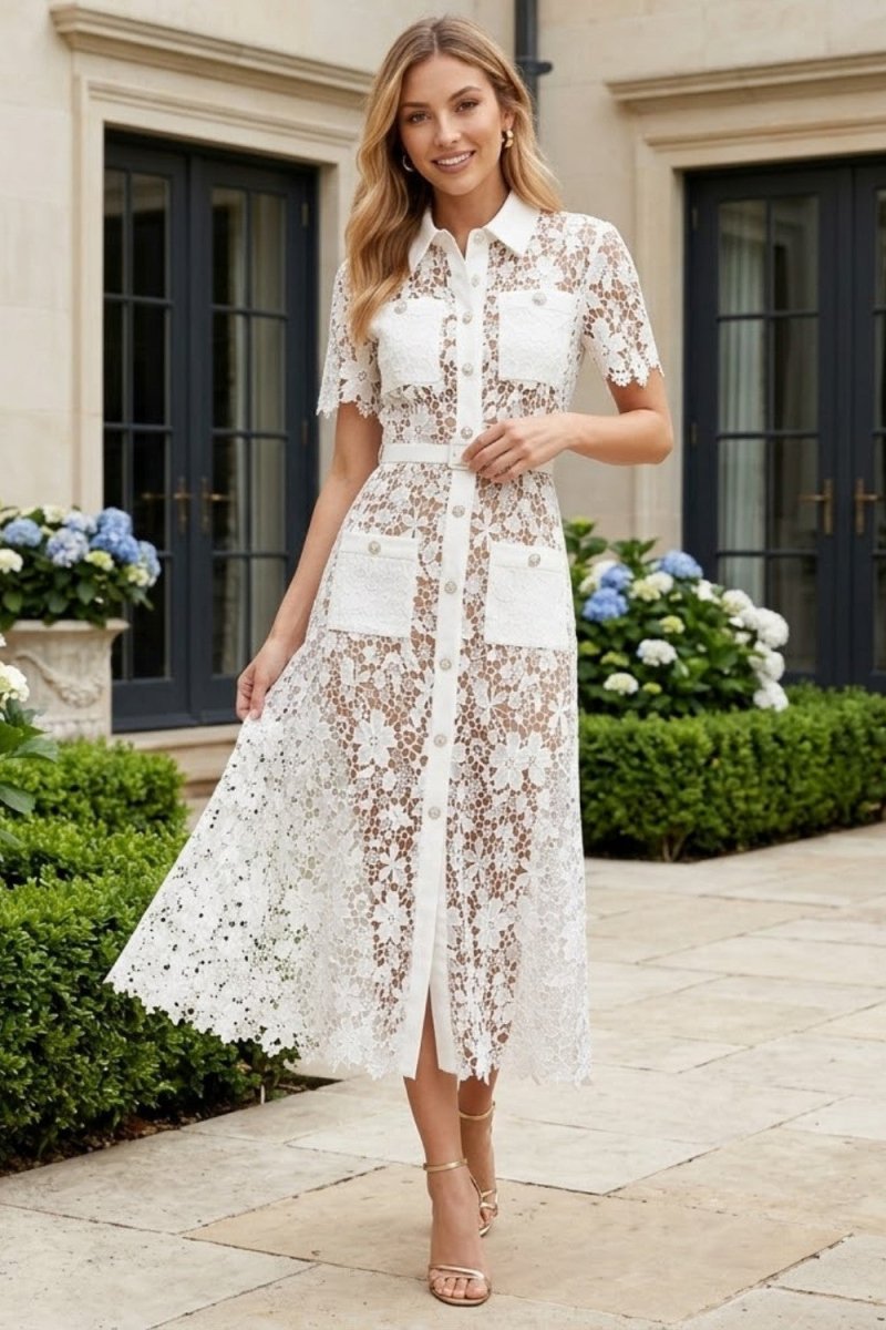 A woman models a white floral lace midi shirt dress with short sleeves, a button-up front, and a tie waist, set against a background of a stone building and green hedges.