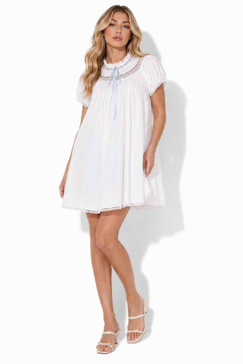 Model wearing Kouvr white cotton babydoll mini dress with lace trim and blue ribbon tie, front full body view.