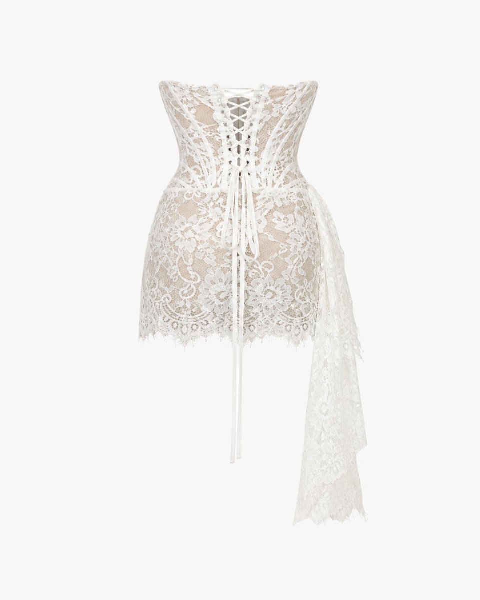 White lace corset-style mini dress with a lace-up back, sheer mesh panels, and a long draped side skirt, shown on a plain light gray background.