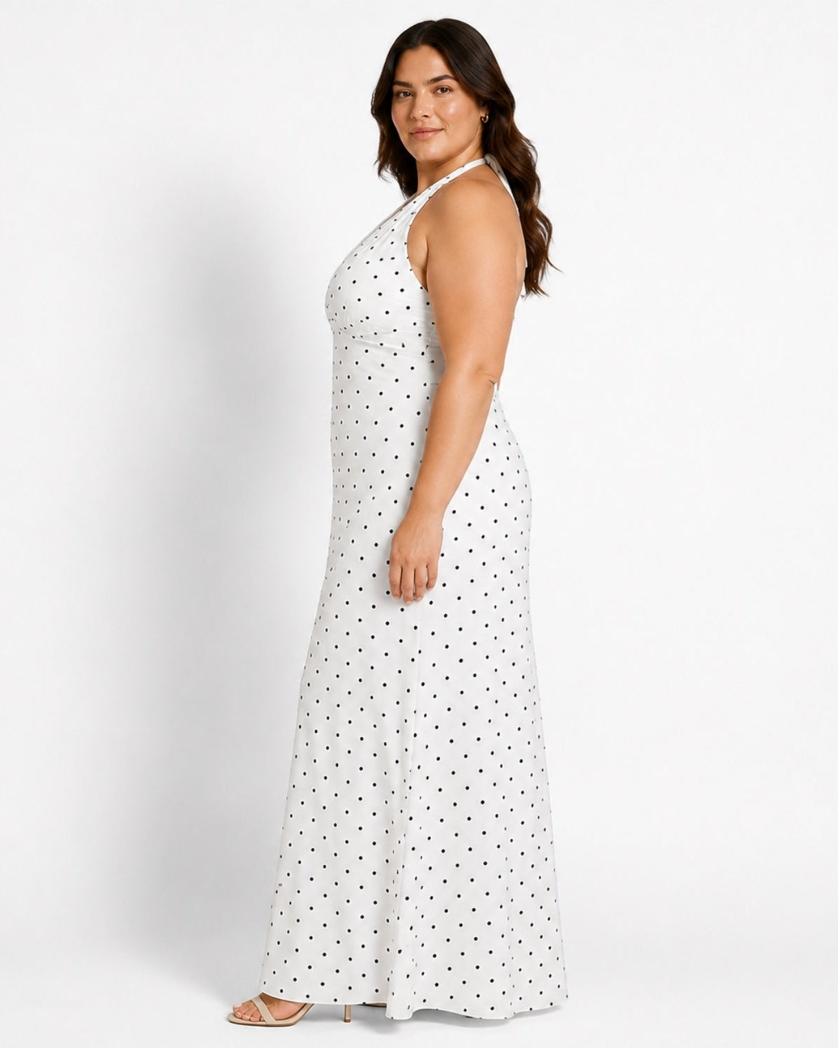 A woman modeling a sleeveless white halter-neck maxi dress with a black polka dot pattern, shown from a side-back angle against a plain white studio background.
