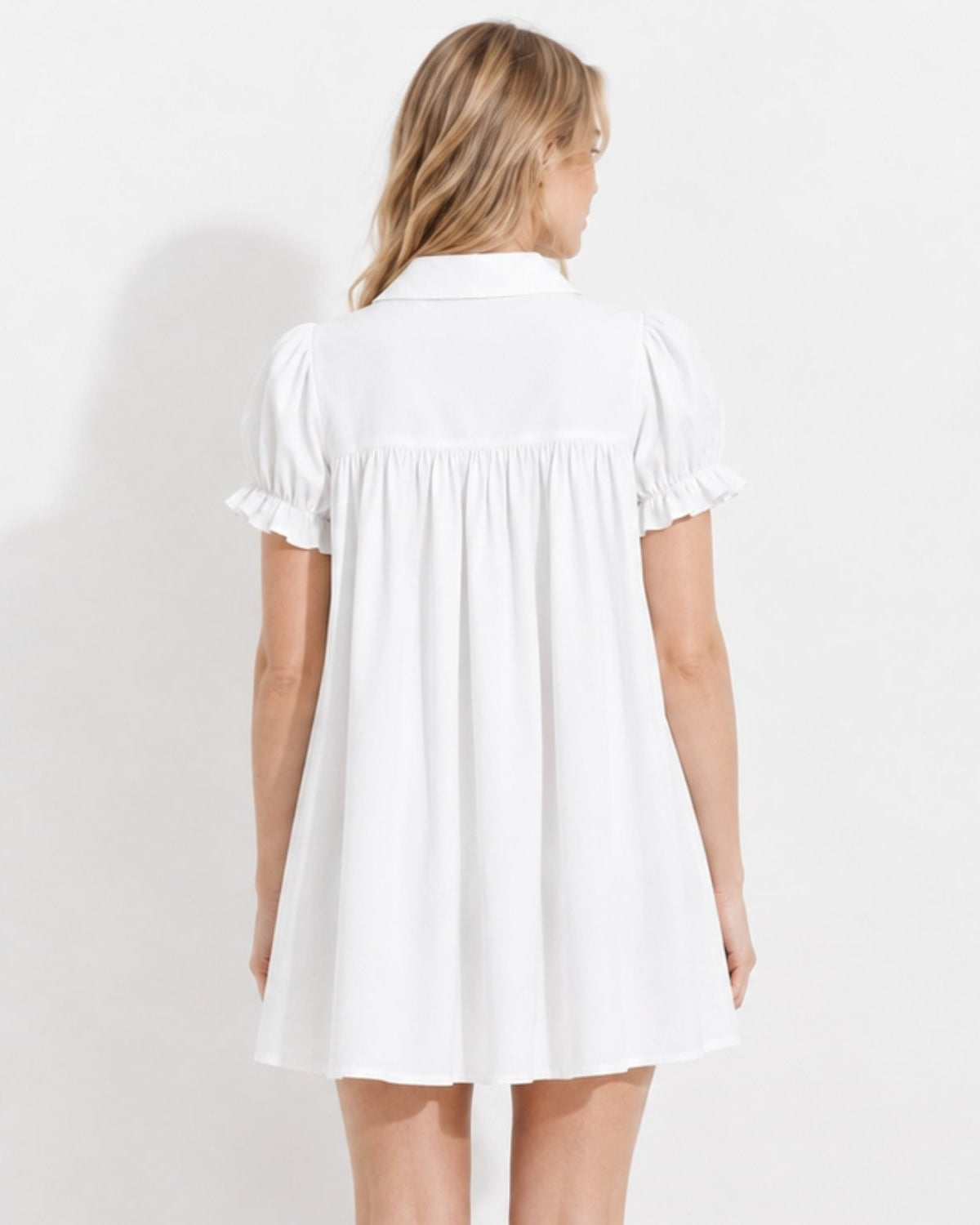 A woman viewed from the back wearing a white short-sleeve dress with ruffled cuffs and a gathered waist seam, photographed against a plain white wall.