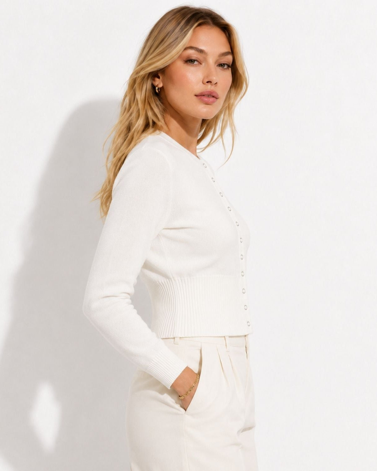 A woman wearing a white ribbed knit cardigan with decorative buttons down the front, paired with cream trousers, standing against a white wall with a soft shadow.