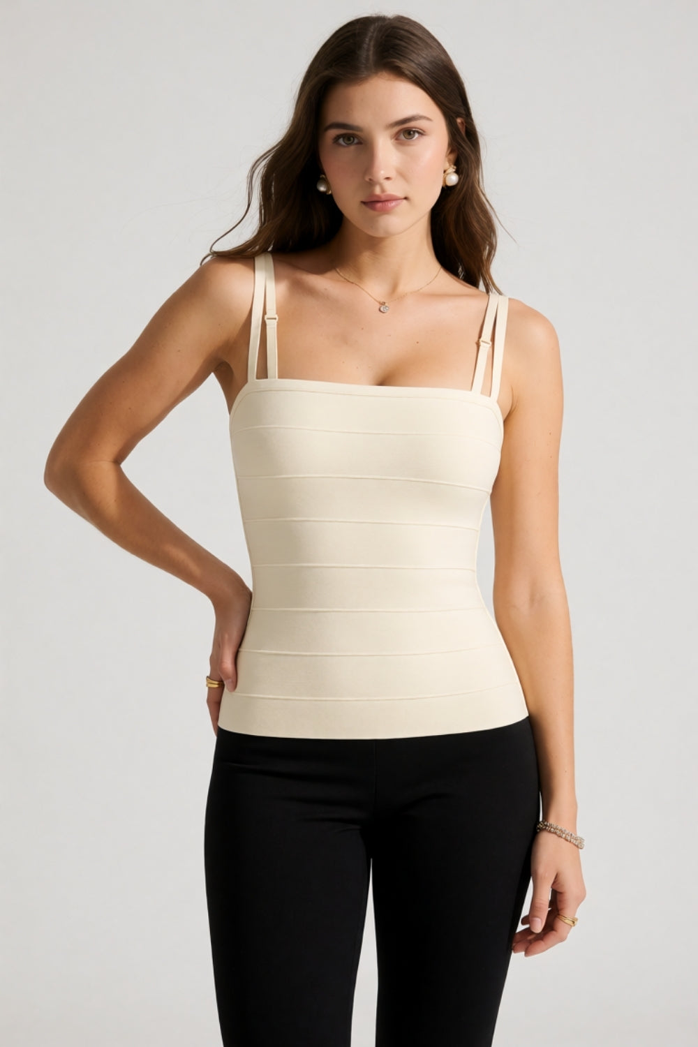 A woman modeling a fitted, white-colored ribbed camisole top with thin shoulder straps and black leggings, photographed against a neutral light gray studio background.