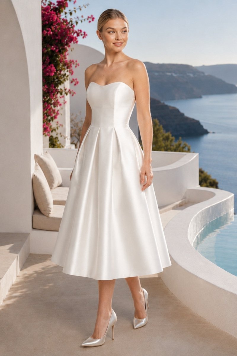 A woman models a strapless white satin A-line midi dress with a sweetheart neckline and a pleated skirt, set against a luxurious outdoor background featuring a pool, white architecture, and a scenic blue sea and mountain vista.