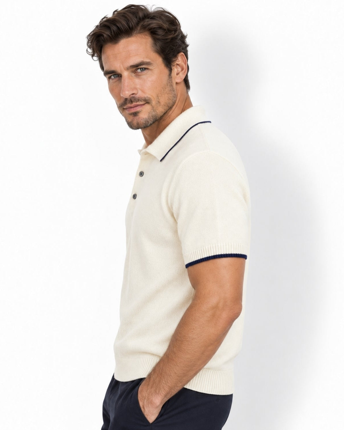 A man wearing a white-colored short-sleeve knit polo shirt with navy blue trim on the collar and sleeve cuffs, paired with dark navy trousers, photographed against a clean white studio background.