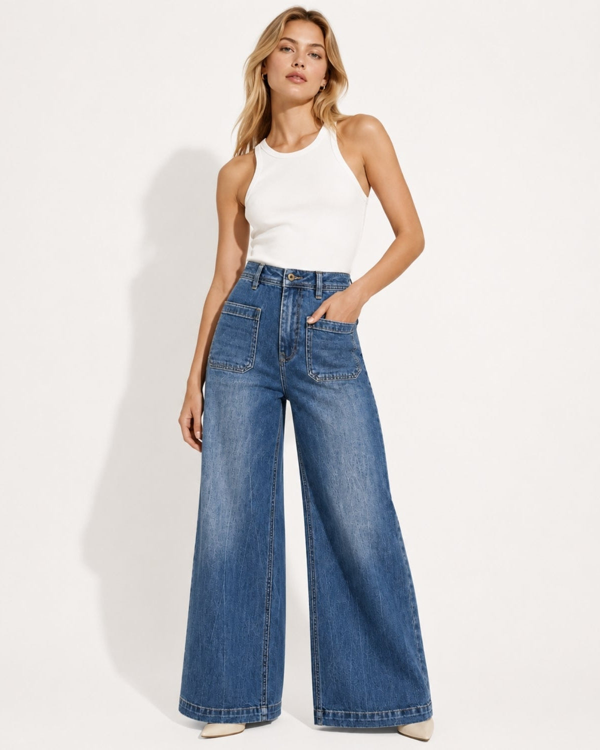 Front lifestyle view of model wearing high-waisted wide leg blue jeans with front patch pockets and a white tank top.