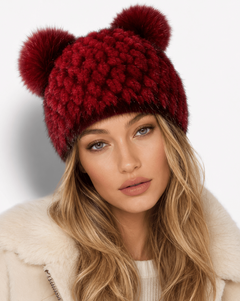 A close-up of a woman wearing a chunky knit beanie with two large pom-poms in deep burgundy red, paired with a cream-colored faux fur coat against a plain white background.
