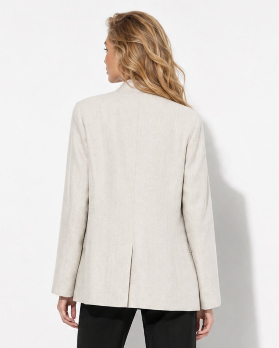 A woman shown from the back wearing a light beige linen blazer and black pants, standing against a plain white studio background.