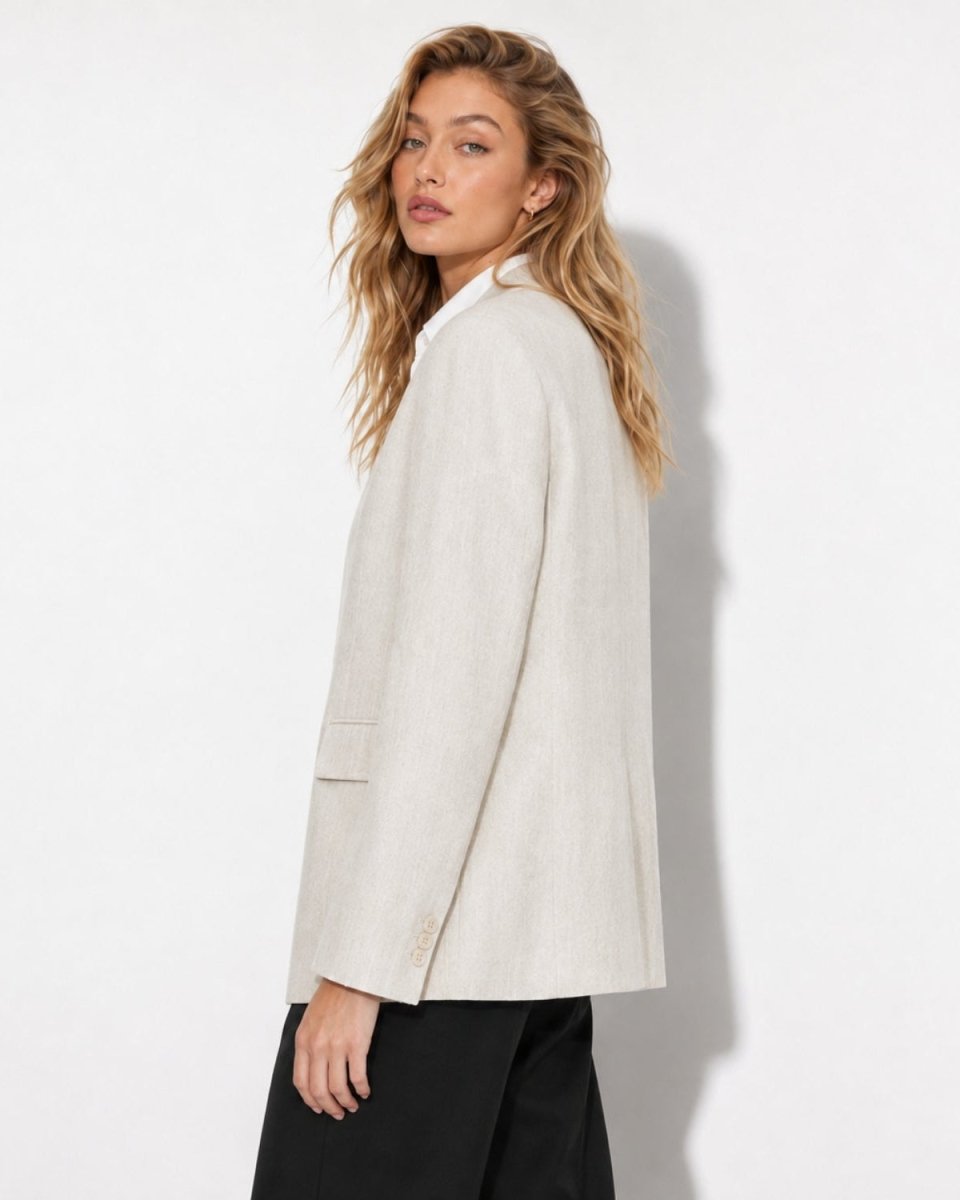 A woman wearing a light beige linen blazer and black trousers, photographed from a side-back angle against a clean white studio background.