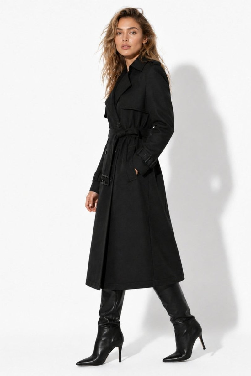 Side view of woman wearing a black belted longline trench coat with pockets