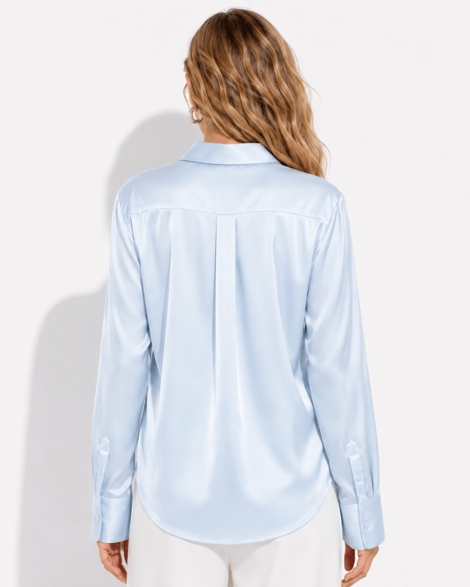 A woman viewed from the back wearing a light blue satin long-sleeve button-up shirt and white pants, set against a plain white studio background.
