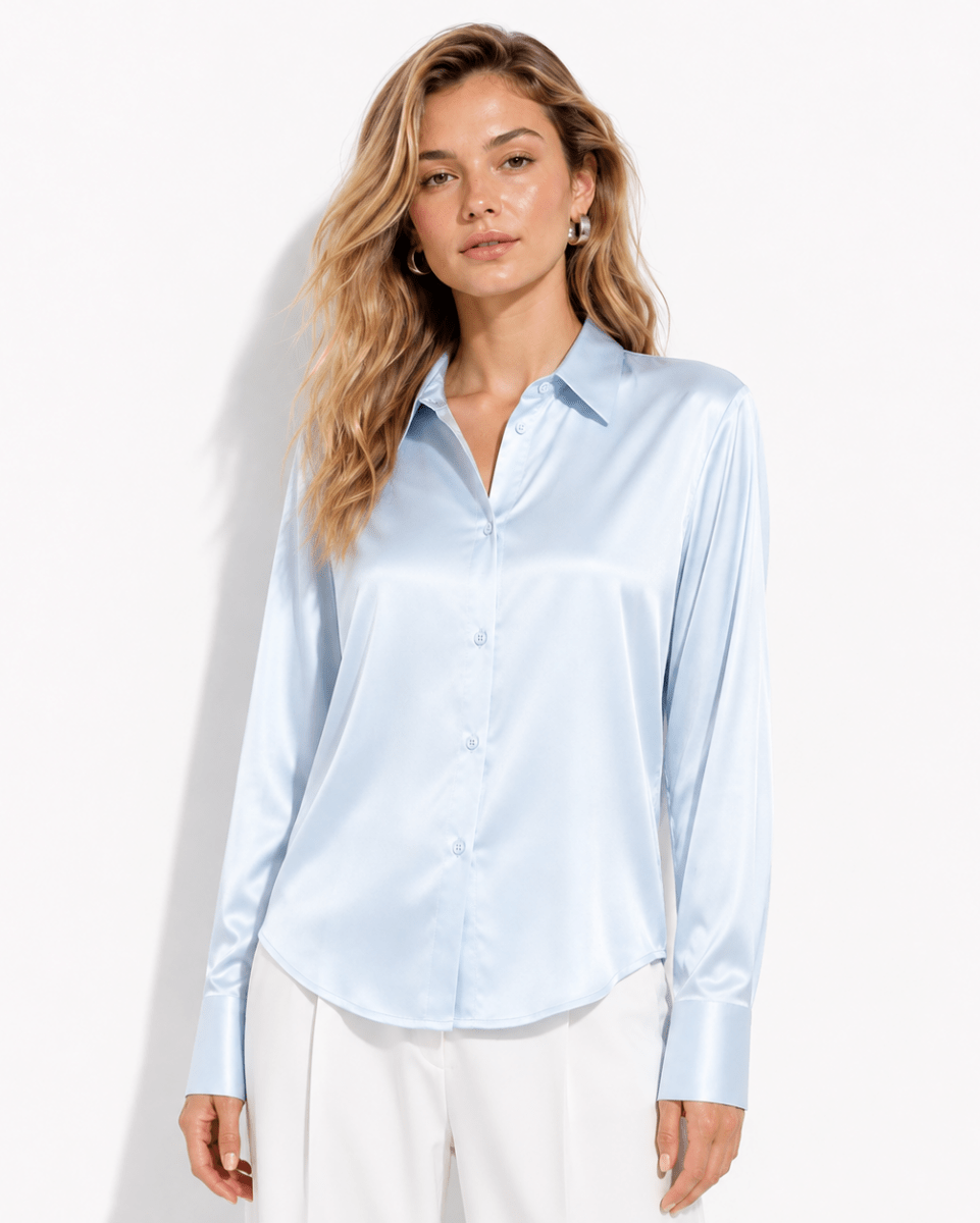 A woman modeling a light blue satin button-up blouse with white trousers, standing against a plain white studio background.