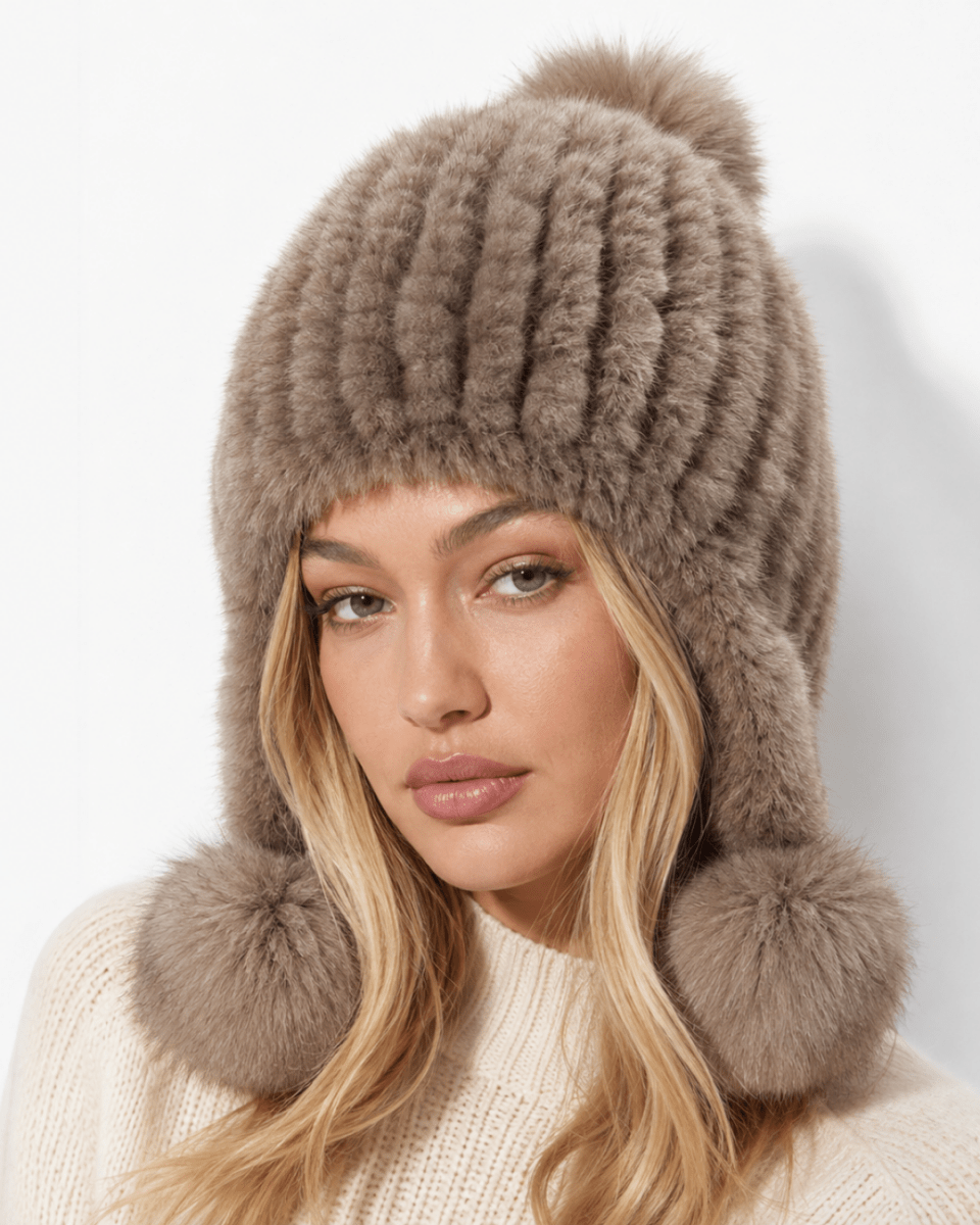 A woman wearing a ribbed faux fur winter hat with ear flaps and large pom-poms in a taupe-brown color, paired with a cream knit sweater against a plain white background.