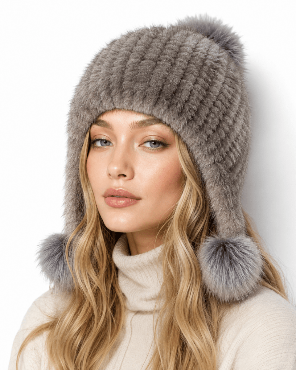 A woman wearing a chunky knit winter beanie in taupe gray with ear flaps and two large pom-poms, paired with a cream turtleneck sweater against a plain white background.