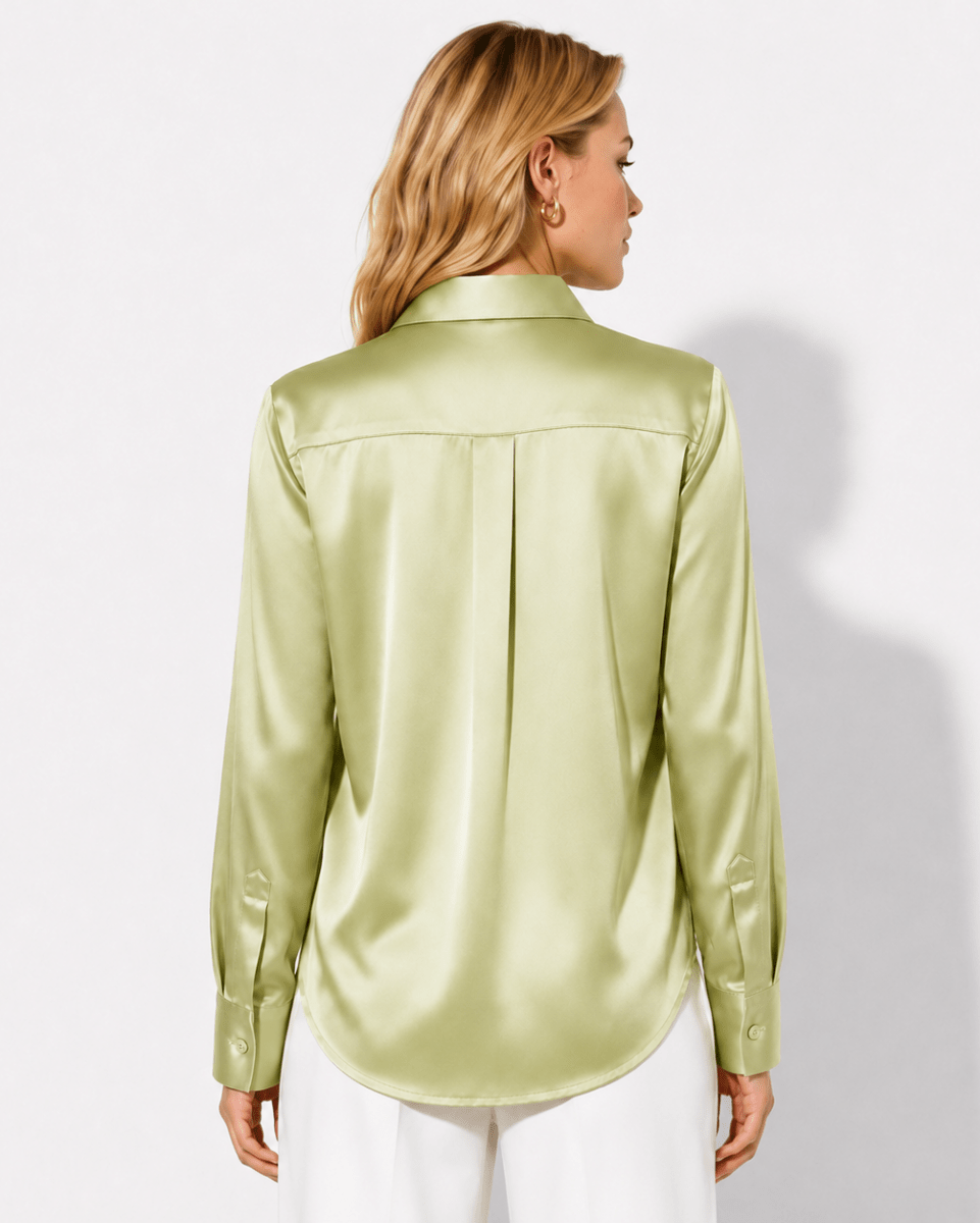 A woman shown from the back wearing a light olive green satin button-down shirt and white pants, photographed against a plain light gray studio background.