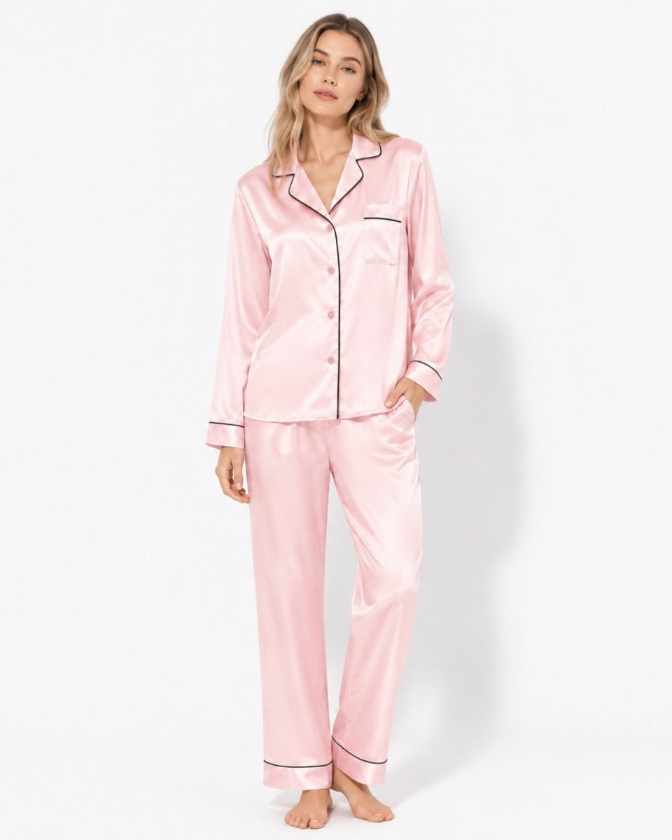 Woman modeling a luxury light pink silk satin pajama set with black contrast piping, long sleeve button down shirt and pants