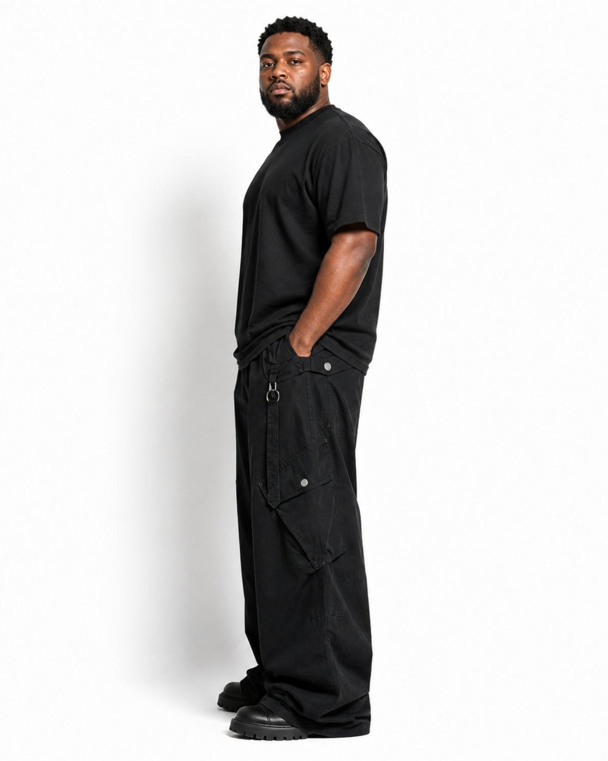 A man wearing a black short-sleeve t-shirt and black wide-leg cargo pants with side utility pockets, standing in profile against a plain white studio background.
