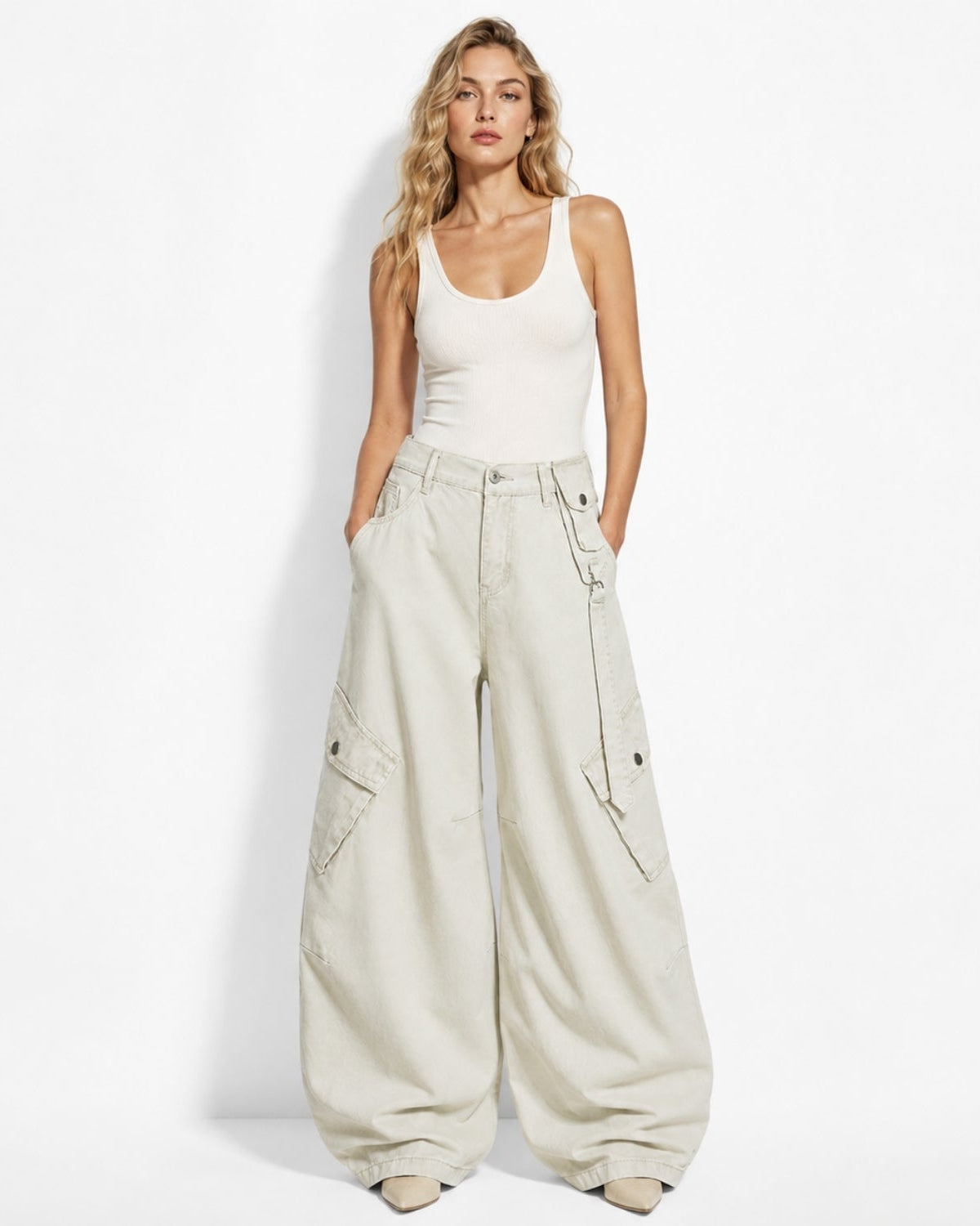 A woman modeling a pair of oversized, wide-leg cargo pants in a light beige color, paired with a white ribbed tank top, standing against a plain white studio background.