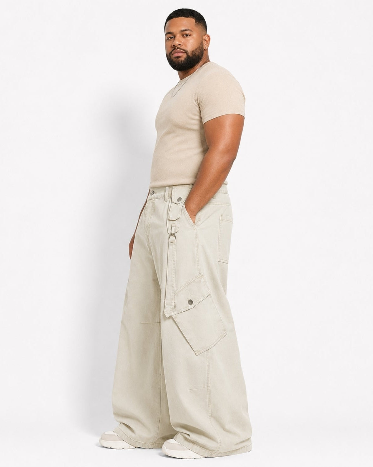 A man wearing a beige short-sleeve t-shirt and off-white wide-leg cargo pants, standing in profile against a clean white studio background.