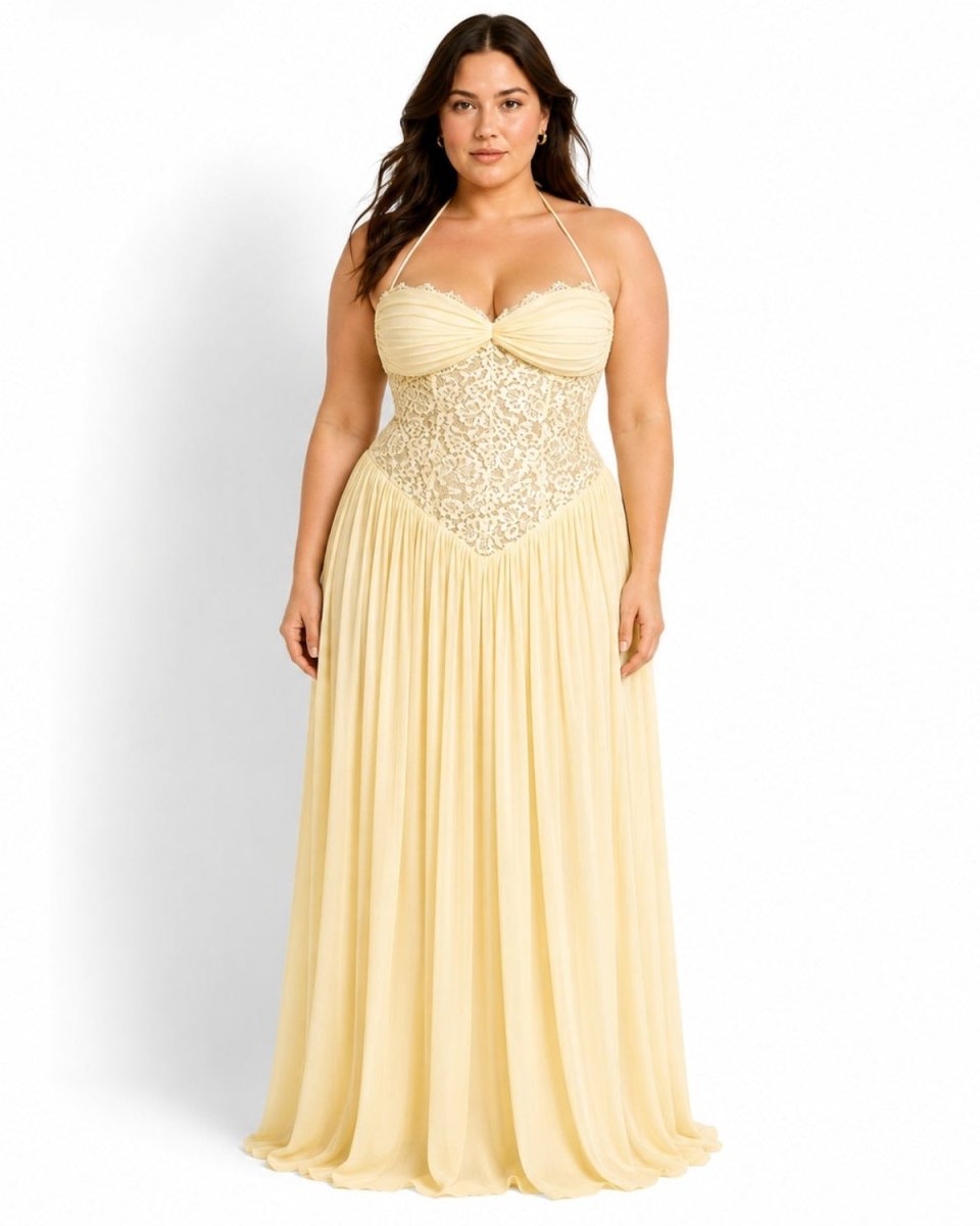 A woman modeling a floor-length cream or pale yellow formal gown with a ruched sweetheart bodice, lace-like embroidered waist panel, thin shoulder straps, and a flowing pleated skirt against a plain white studio background.
