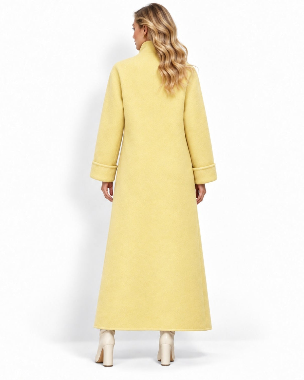 A woman viewed from the back wearing a long, light yellow coat with a high collar and wide sleeves, paired with cream high-heeled boots, against a plain white studio background.