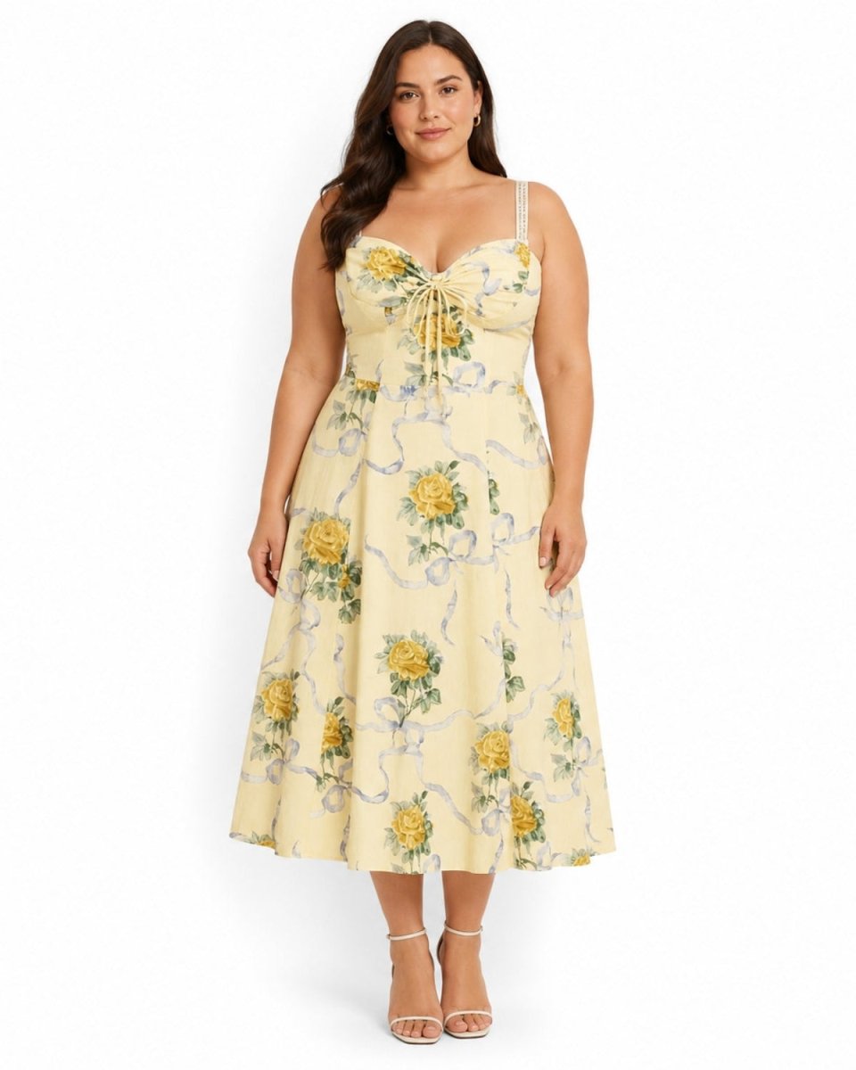 A woman modeling a light yellow midi dress with a yellow and green floral print, featuring a sweetheart neckline, thin straps, and a tie detail at the bust. She is wearing clear high-heeled sandals against a plain white background.